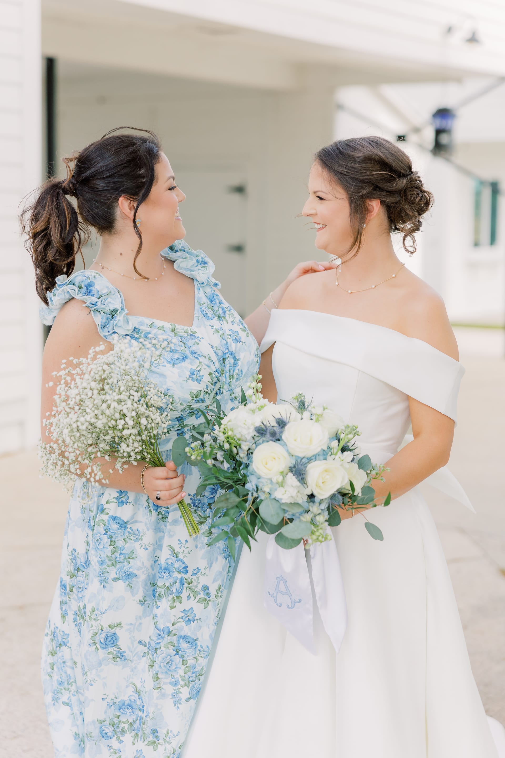 Bride in an off-the-shoulder white gown holding a blue and white bouquet smiles with her bridesmaid in a blue floral dress holding baby's breath outside Scarborough House.