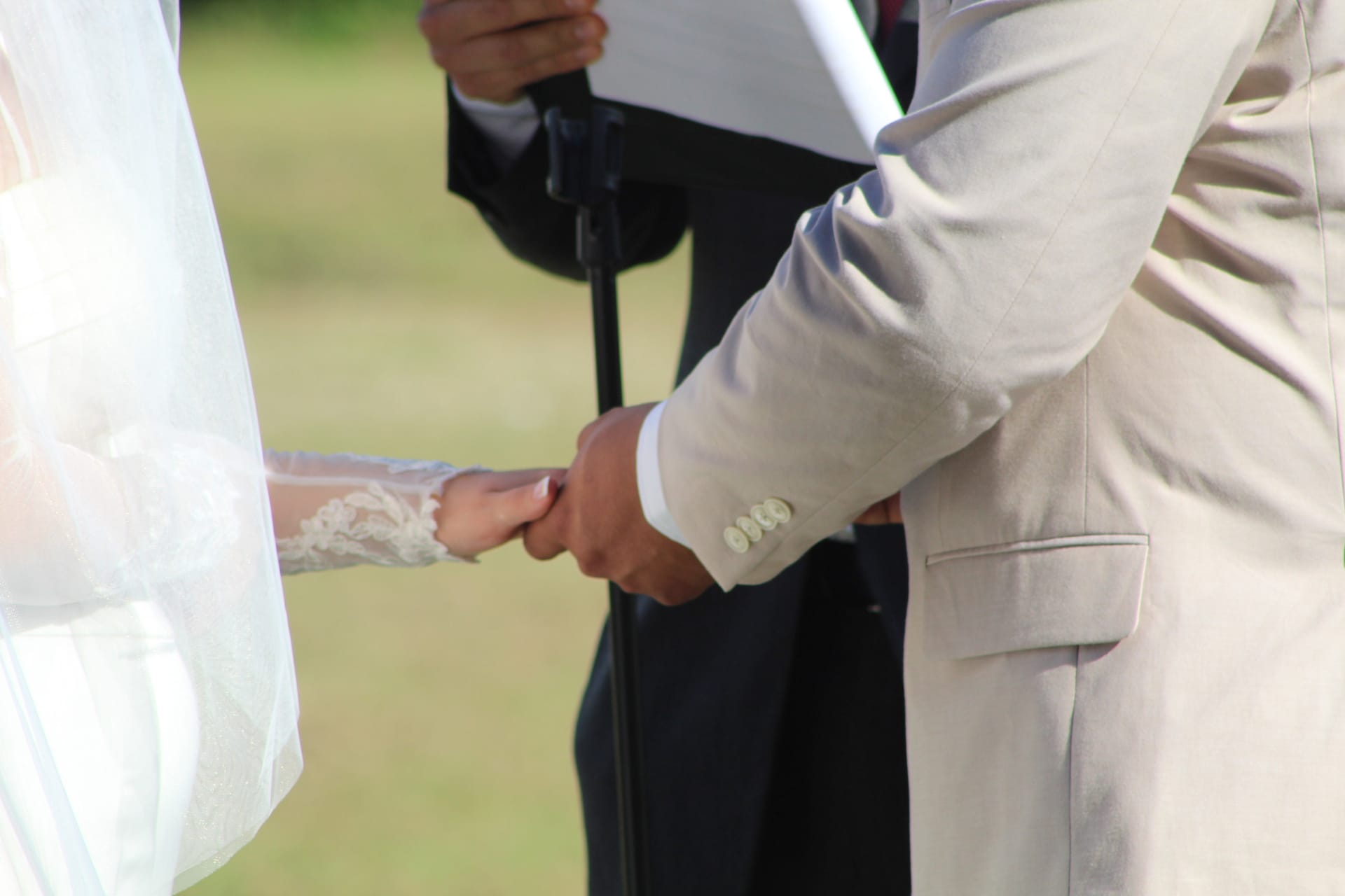 Bride in lace-sleeved gown and white veil holds hands with groom in a tan suit during an outdoor wedding ceremony at Scarborough House while the officiant reads from a book in the background.