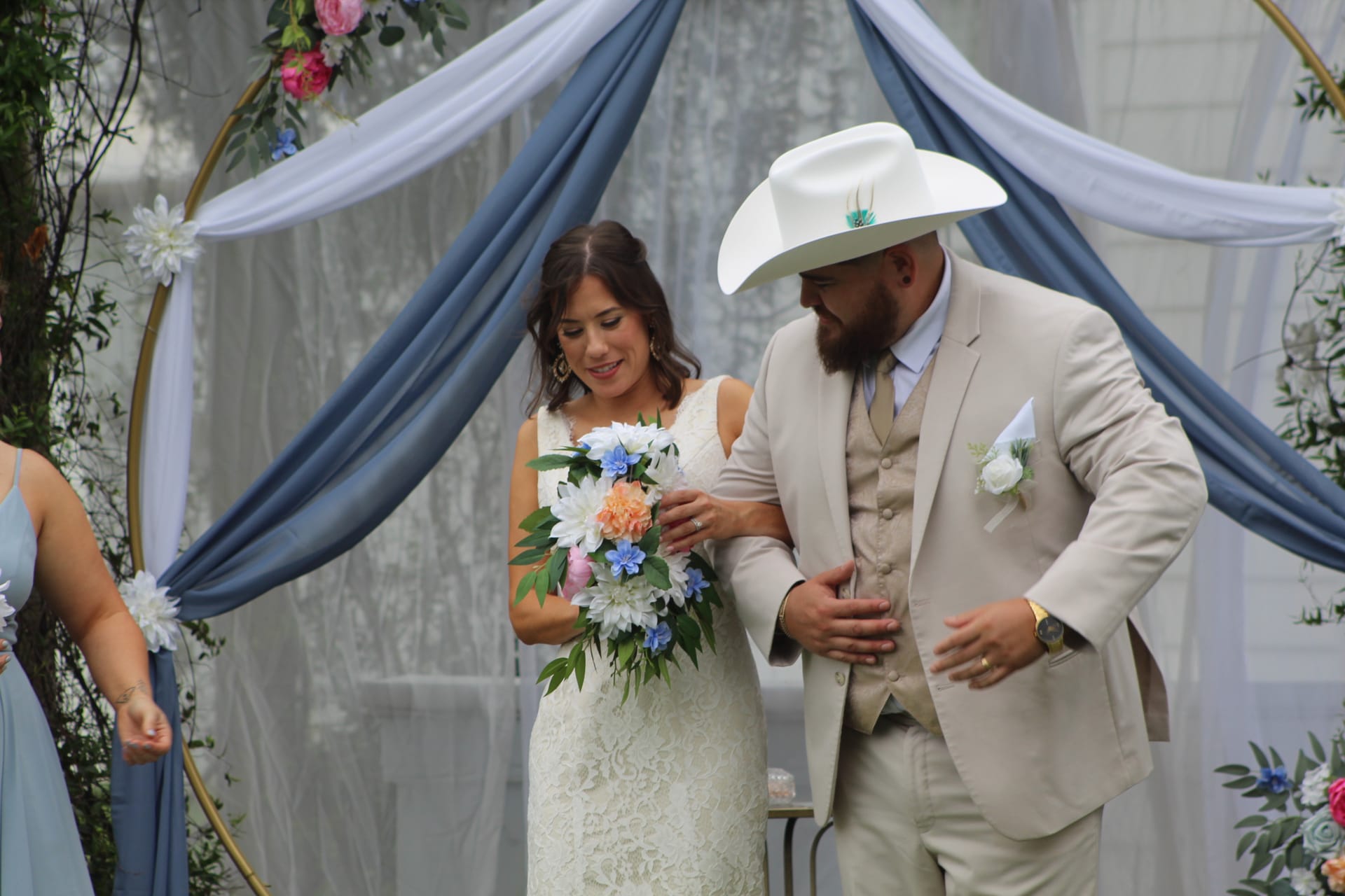 Bride in a lace gown holding a colorful bouquet stands beside a bearded groom in a tan suit and white cowboy hat beneath a floral-draped circular arch at an outdoor wedding ceremony.