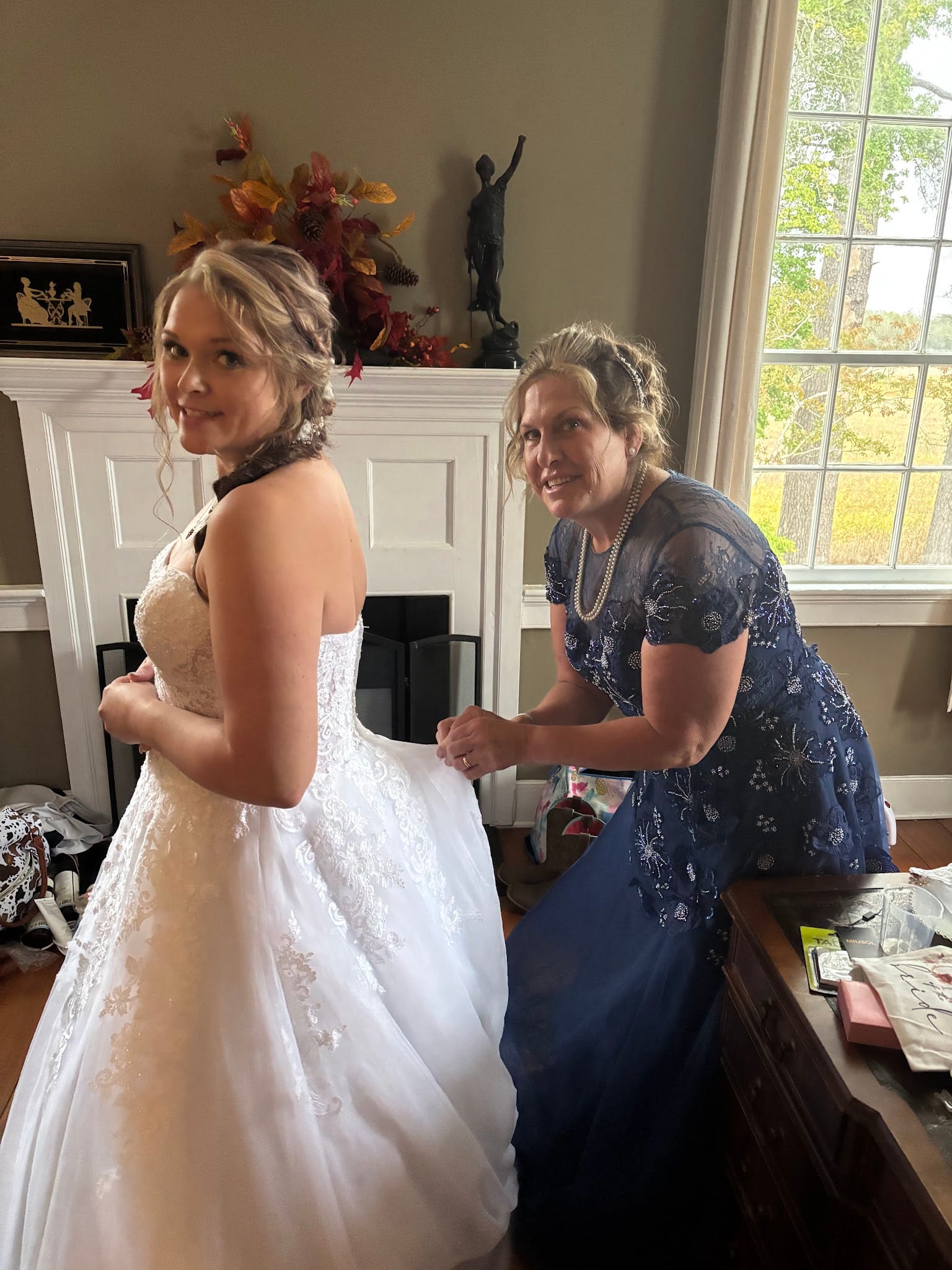 A bride in a white lace ball gown looks over her shoulder smiling while a woman in a navy beaded dress fastens the back of the dress in a warmly decorated room.