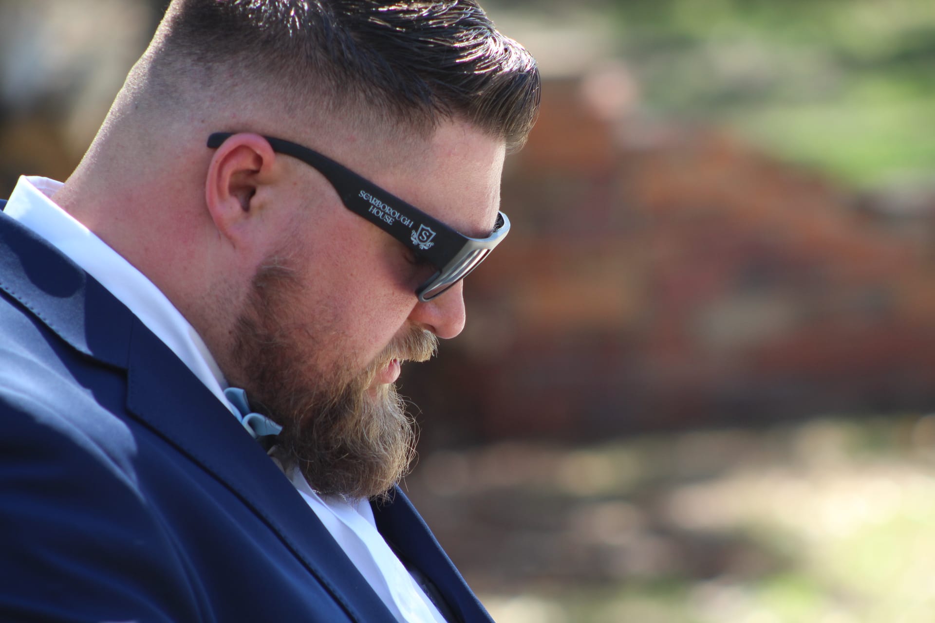 A bearded groom in a navy suit and Scarborough House sunglasses gazes downward in a thoughtful moment before the ceremony at Scarborough House wedding venue.