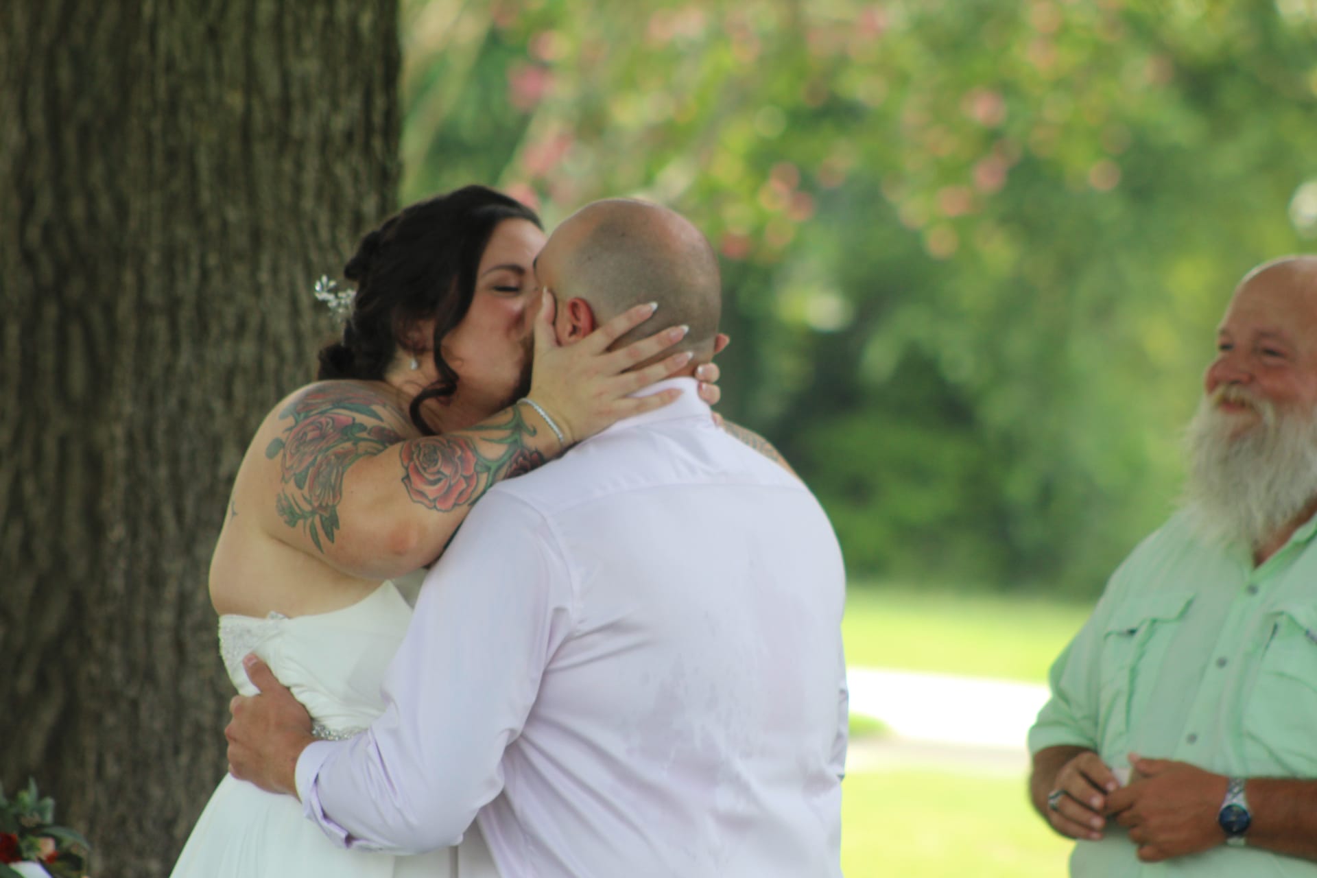 Bride with floral arm tattoos in a white wedding gown shares her first kiss with the groom beneath a large oak tree as a bearded officiant smiles warmly beside them at an outdoor ceremony.