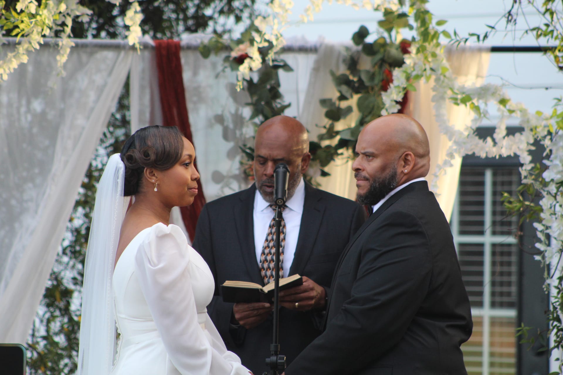 Bride in a white long-sleeve gown and veil exchanges vows with her tuxedo-clad groom before an officiant reading from a book beneath a floral-draped arch at Scarborough House.