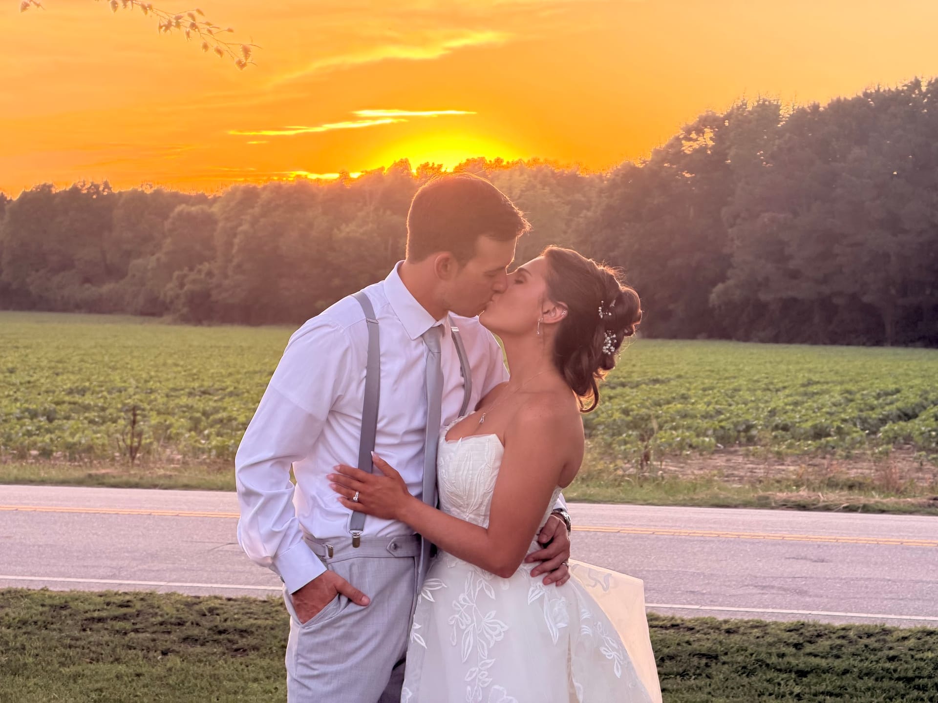 Bride and groom sharing a kiss in front of a glowing orange sunset sky with open farmland fields and trees in the background at Scarborough House.