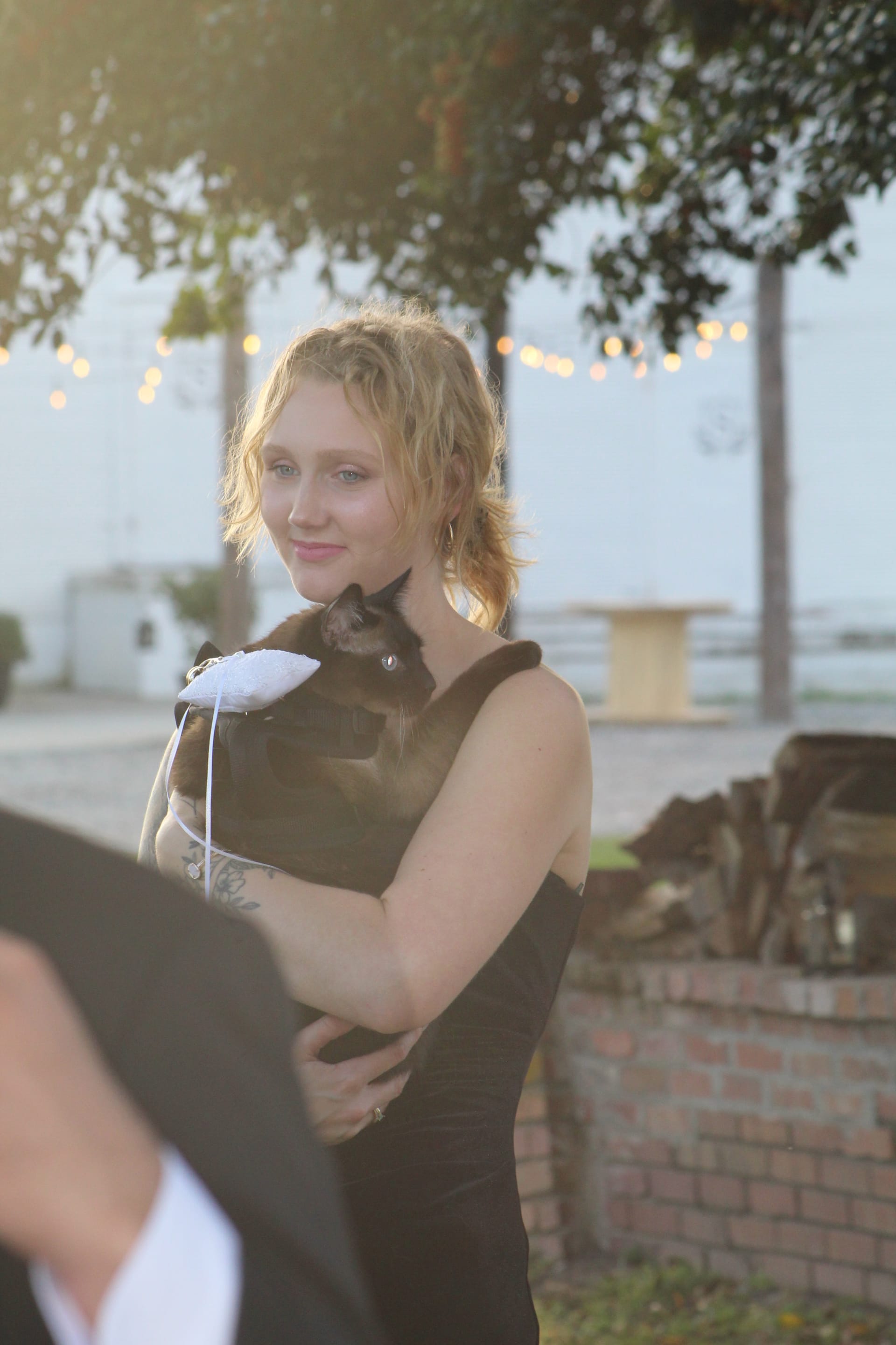 A smiling bridesmaid in a black dress holds a Siamese cat wearing a small white ring-bearer pillow during an outdoor golden-hour wedding celebration.