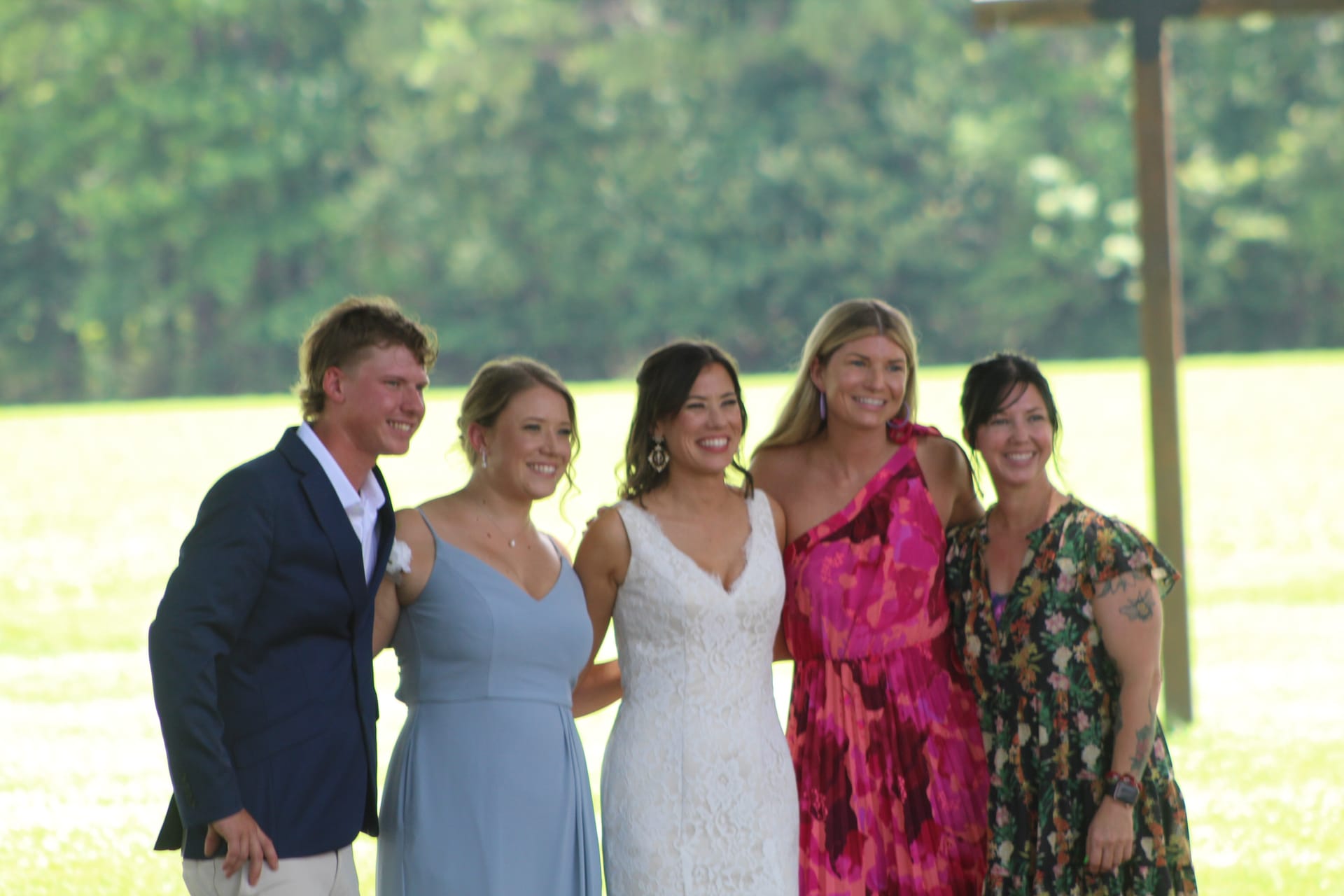 Bride in white lace gown laughing with four guests including a bridesmaid in dusty blue and a woman in a vibrant pink dress on the sunlit ceremony lawn surrounded by lush greenery.
