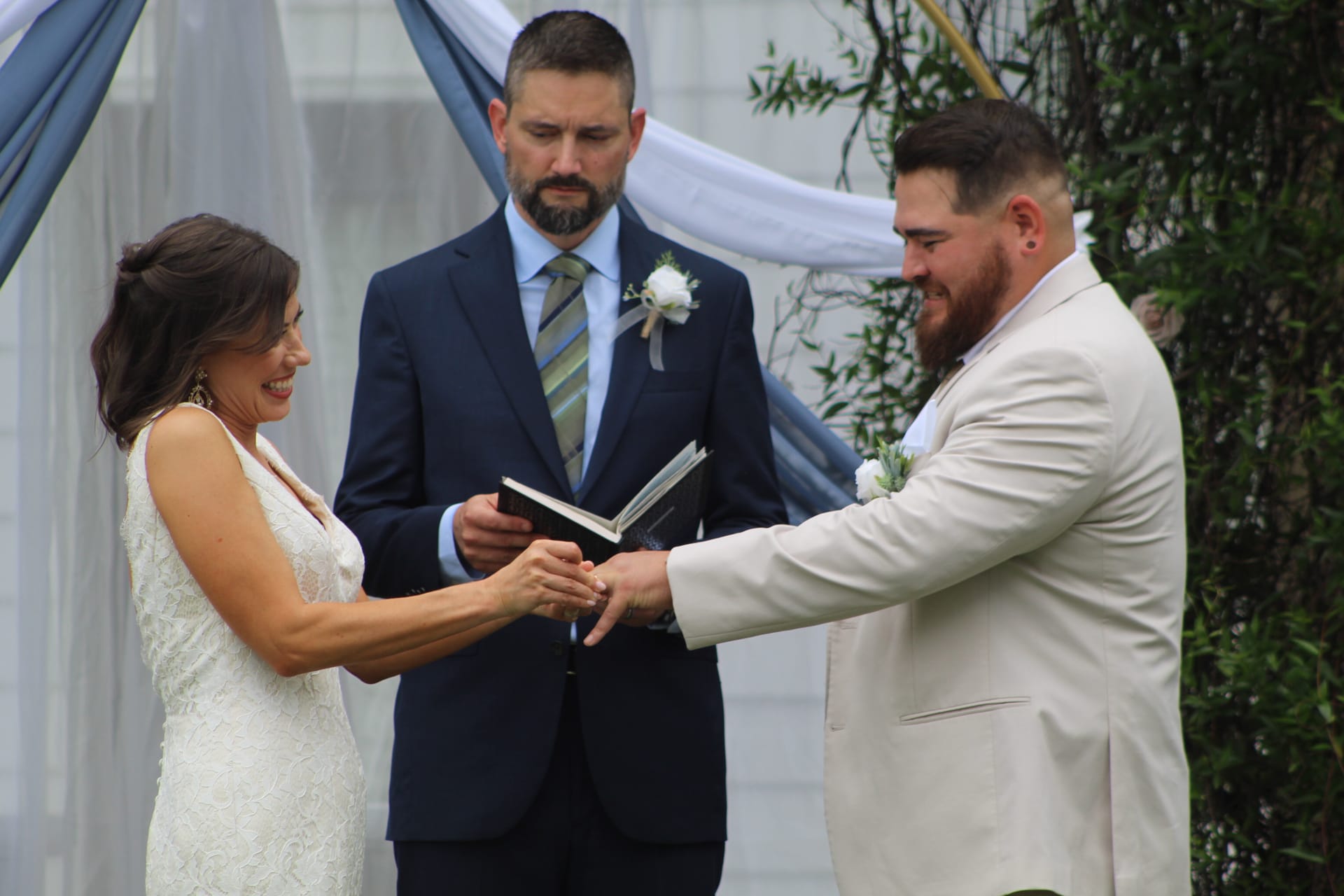 Bride and groom exchange rings while smiling during their outdoor wedding ceremony at Scarborough House, with the officiant holding a book beneath a draped blue and white arch.