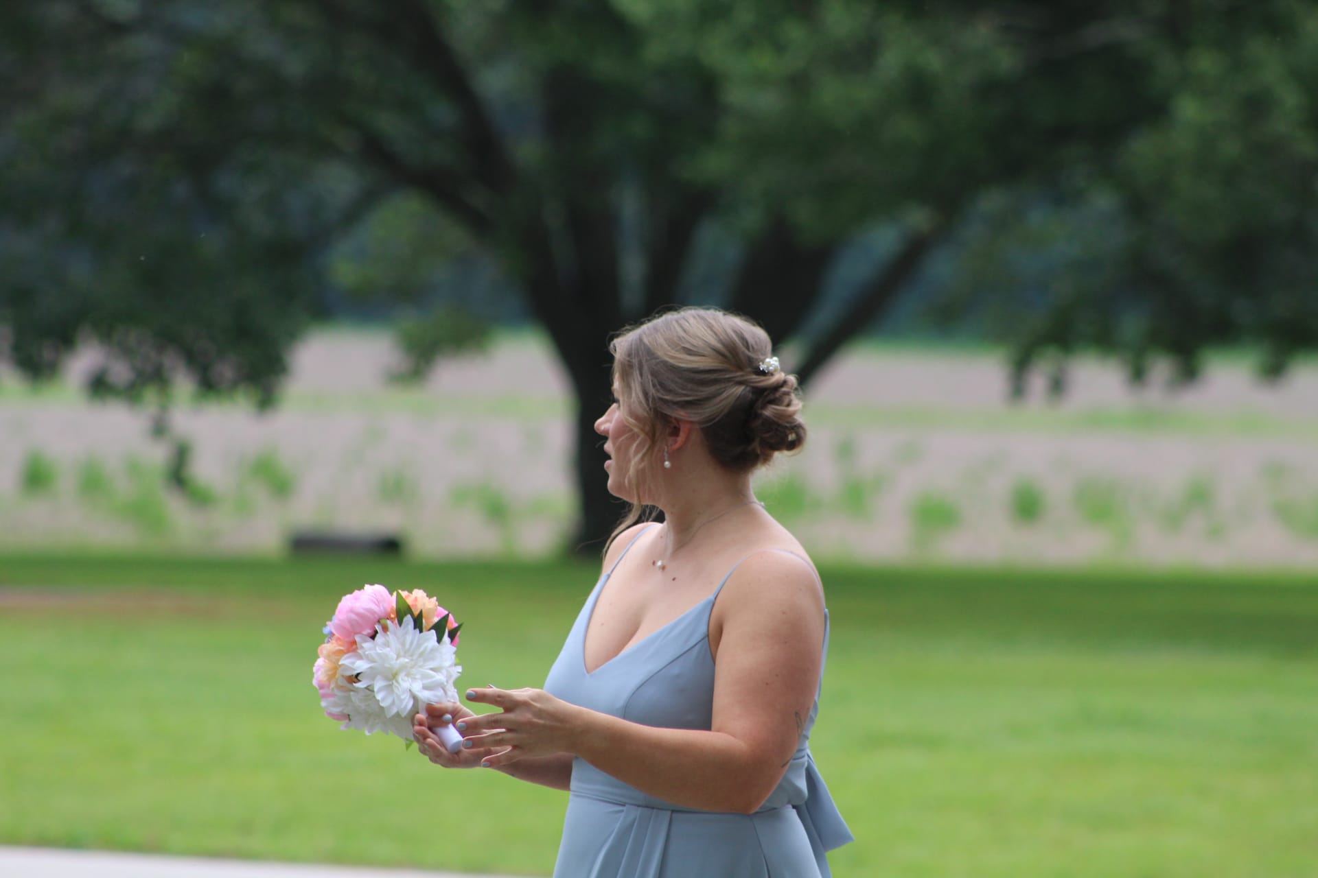 Bridesmaid in a dusty-blue gown holding a pink and white floral bouquet on the ceremony lawn at Scarborough House with grand oak trees in the background.