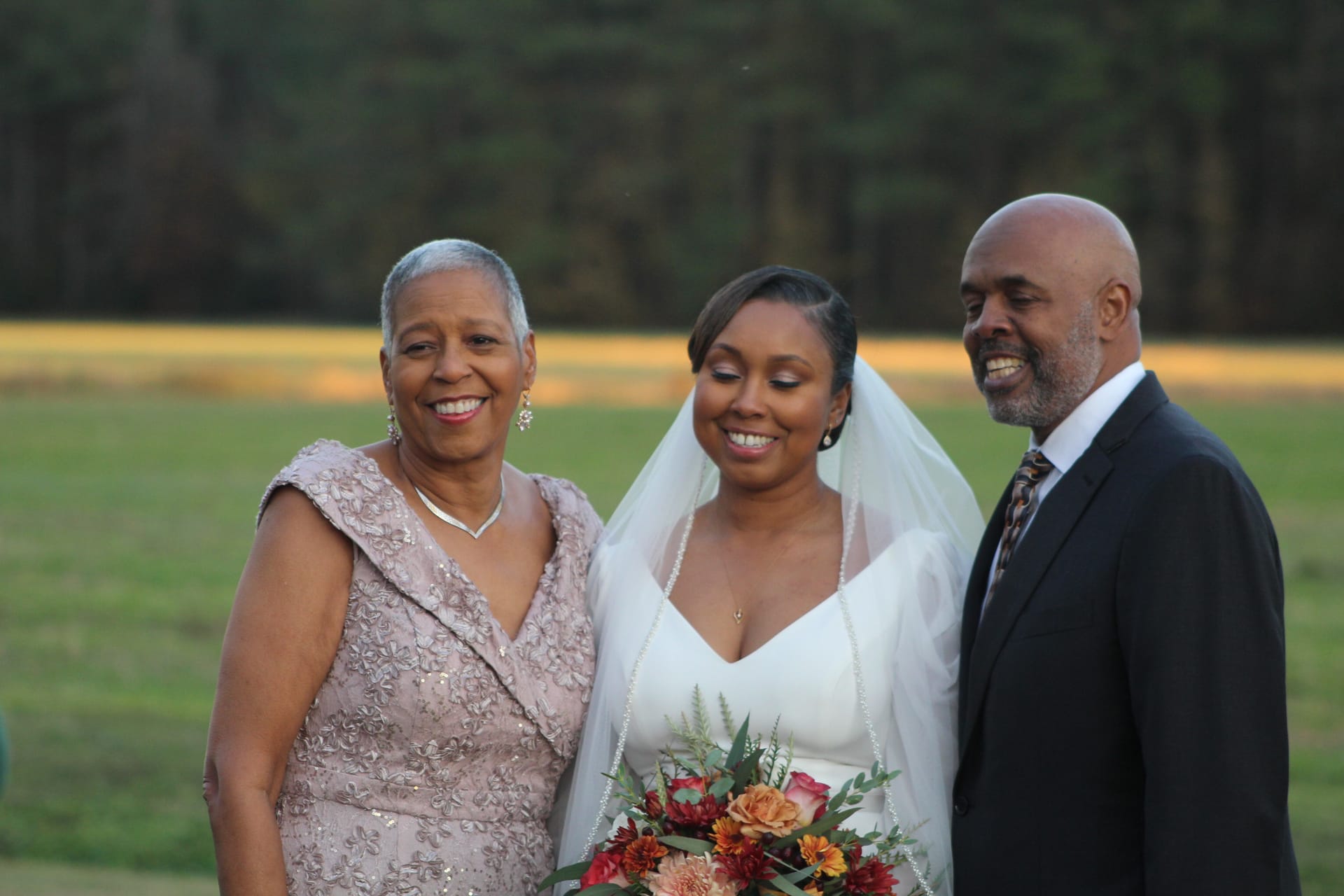 A radiant bride in a white gown and veil holds an autumn bouquet while posing with her smiling parents on the open farmland fields at Scarborough House during golden hour.