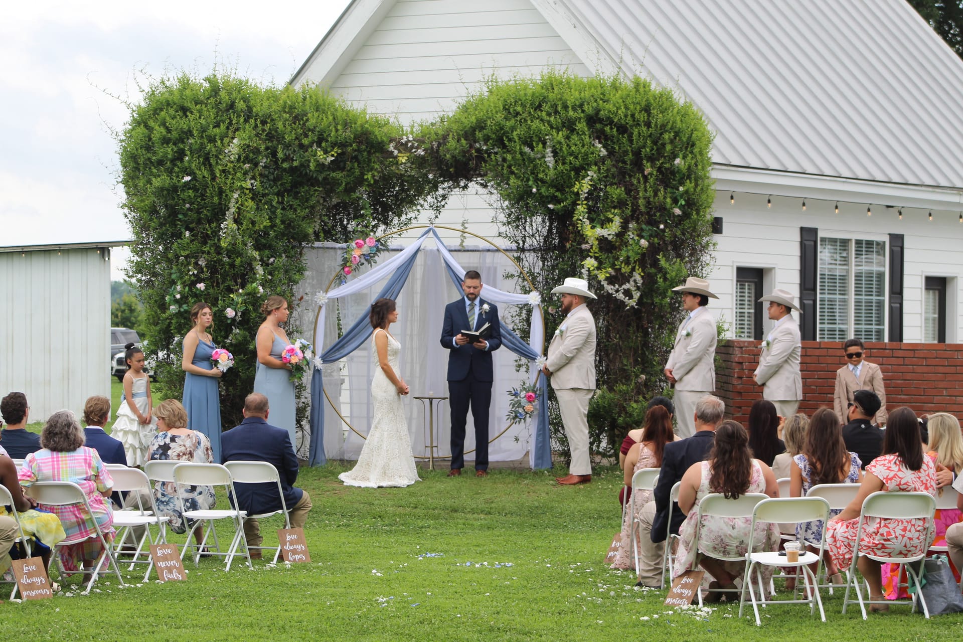 Bride and groom exchange vows beneath a lush greenery and blue-draped arch on the ceremony lawn at Scarborough House, surrounded by bridesmaids, groomsmen, and seated guests.