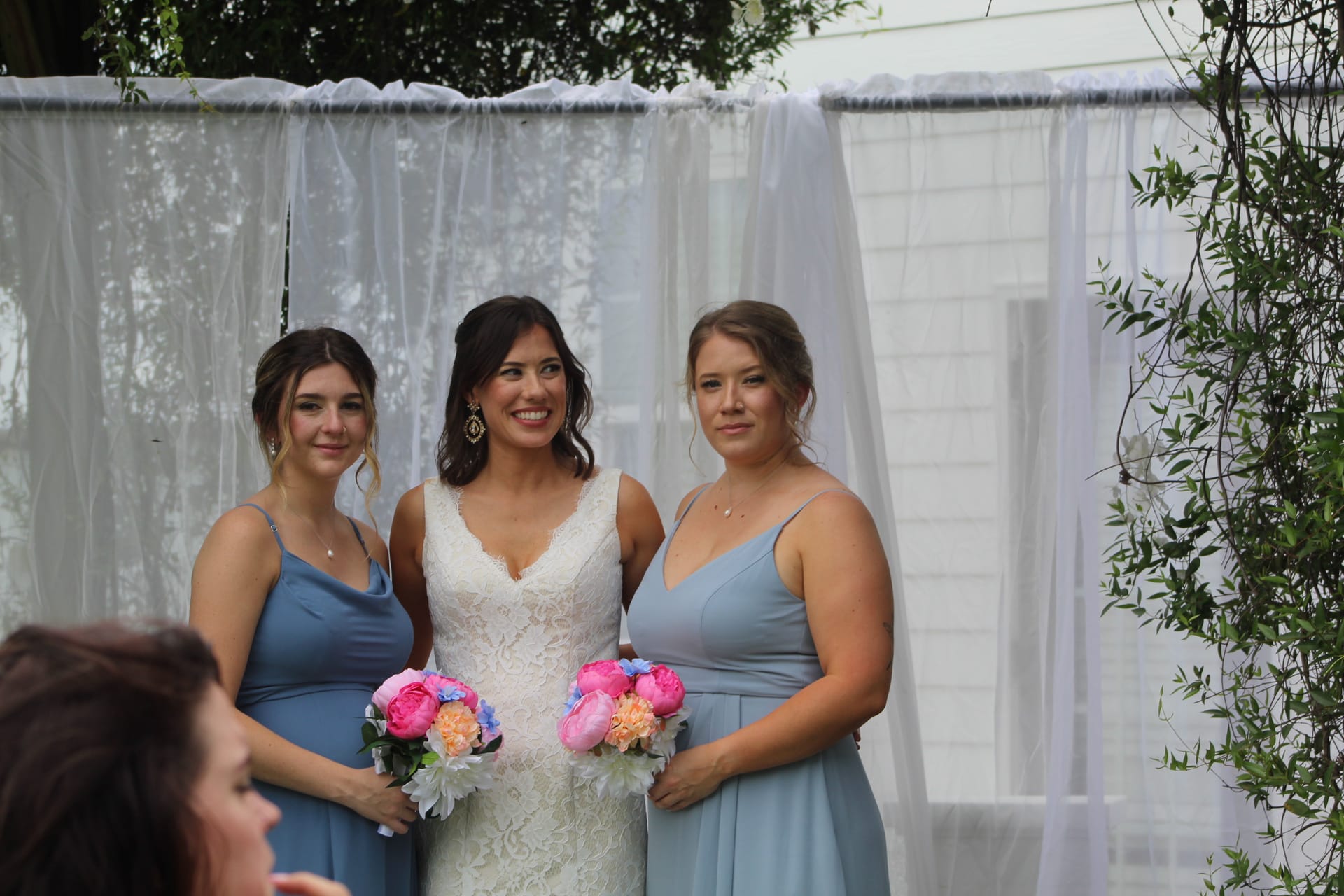Bride in white lace gown stands smiling between two bridesmaids in dusty blue dresses holding pink and peach floral bouquets in front of a white draped ceremony backdrop at Scarborough House.
