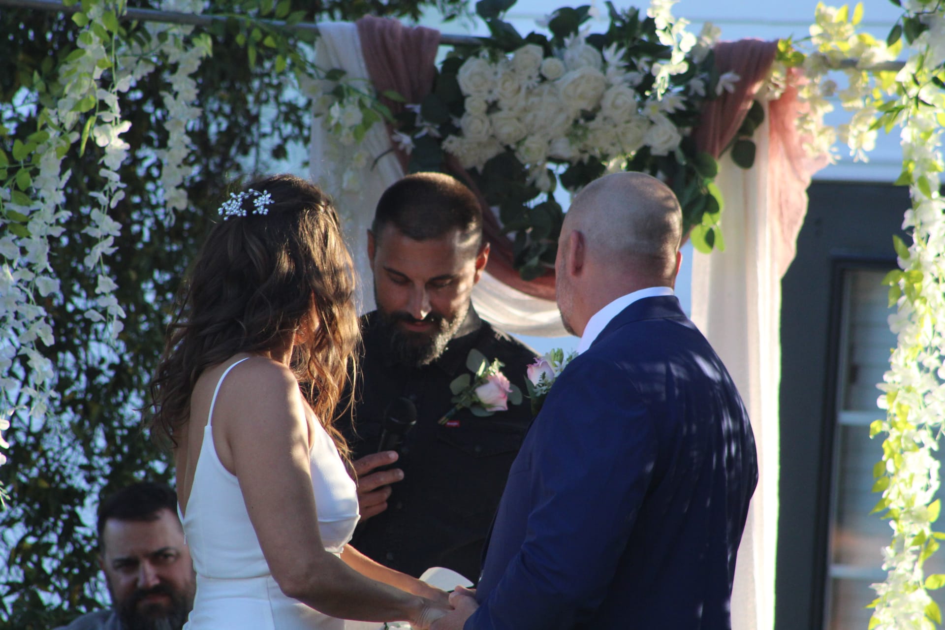 Bride and groom exchange rings beneath a lush floral arch adorned with white roses and greenery during an outdoor wedding ceremony at Scarborough House.
