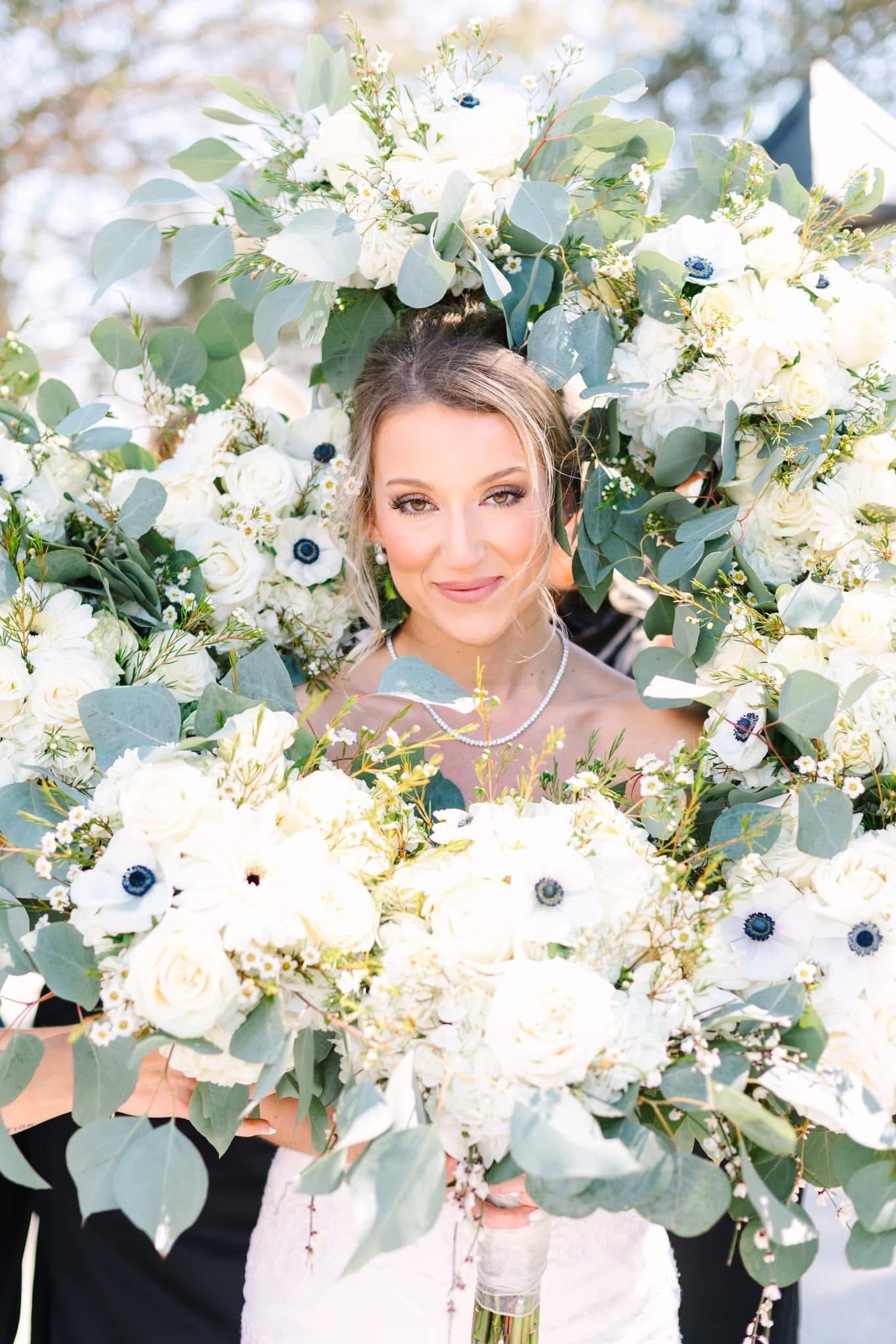 Bride smiling warmly while framed by an arch of white roses anemones and eucalyptus bouquets held by bridesmaids in black dresses at Scarborough House estate wedding venue.