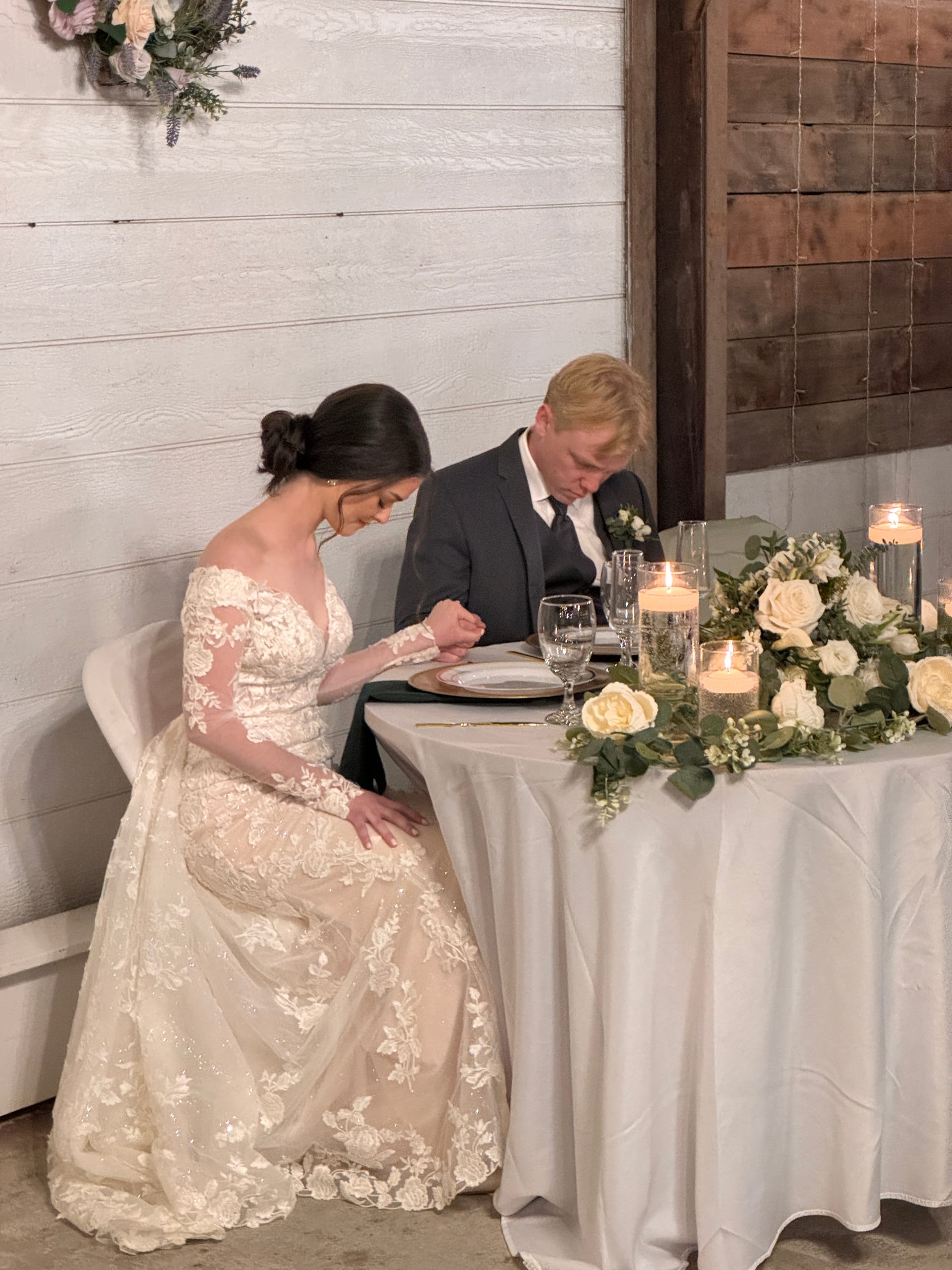 Bride in a long-sleeve lace gown and groom in a dark suit bow their heads in prayer at a candlelit sweetheart table adorned with white roses and greenery at Scarborough House.