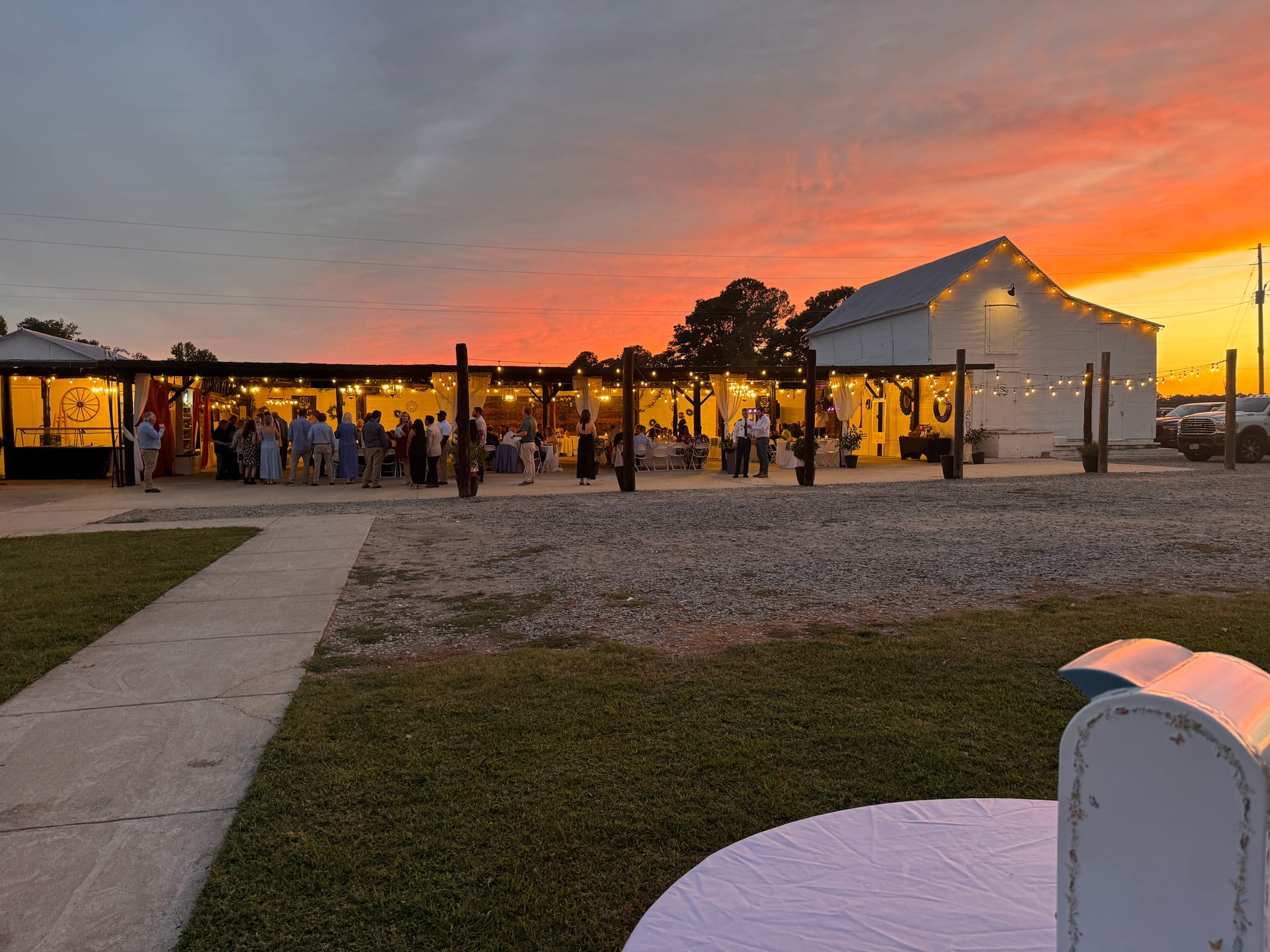 Wedding reception guests mingling under warm string lights at a rustic barn venue bathed in a vivid orange and pink sunset sky.