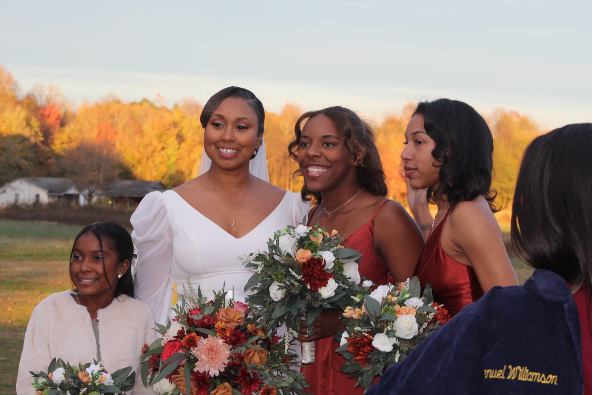 Bride in white gown and bridesmaids in burgundy dresses smile and hold lush autumn floral bouquets during golden hour on the scenic farmland fields at Scarborough House.