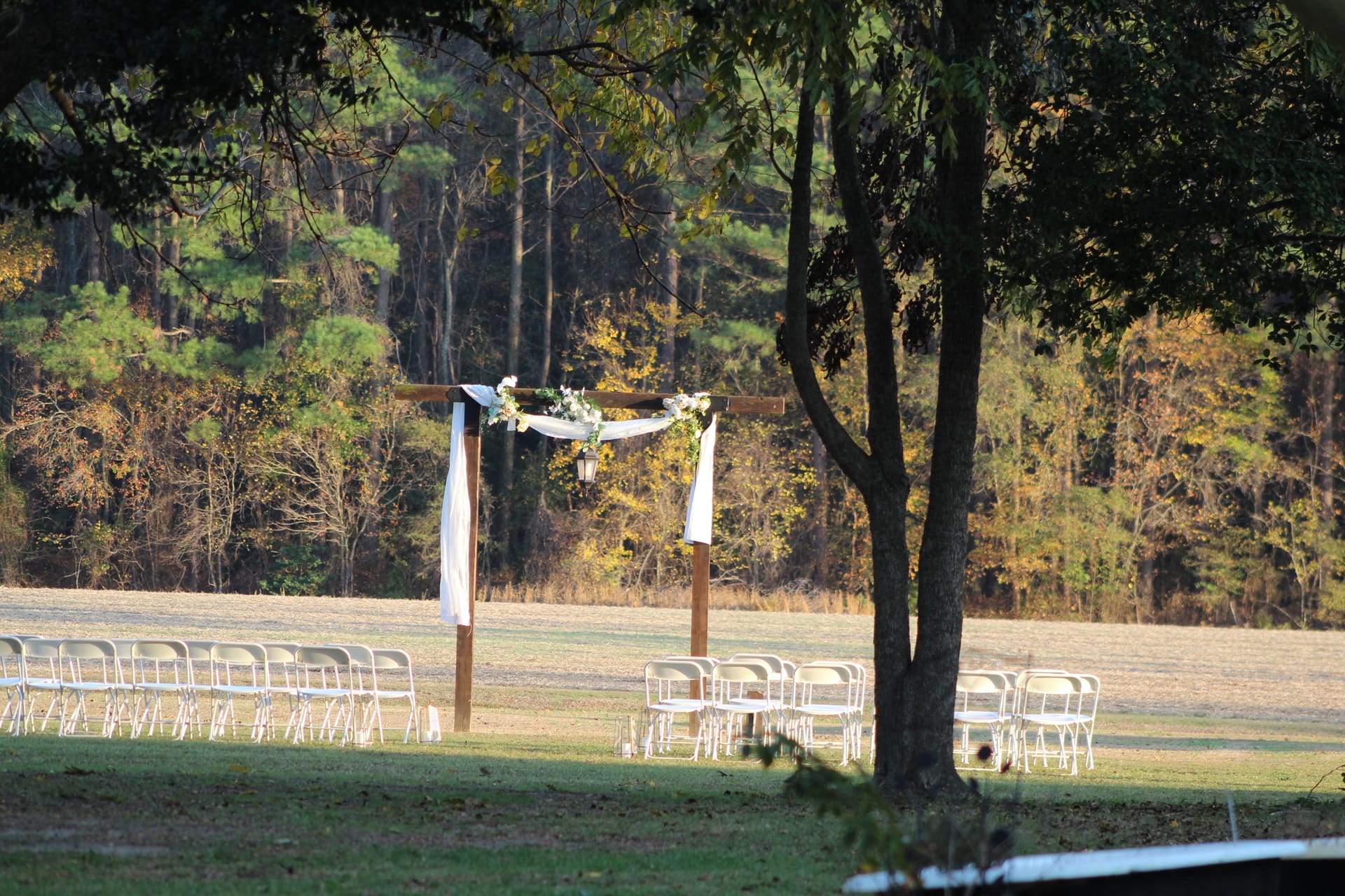 A rustic wooden wedding arch draped in white fabric and florals stands on the open ceremony lawn at Scarborough House, flanked by rows of white folding chairs and framed by autumn trees.