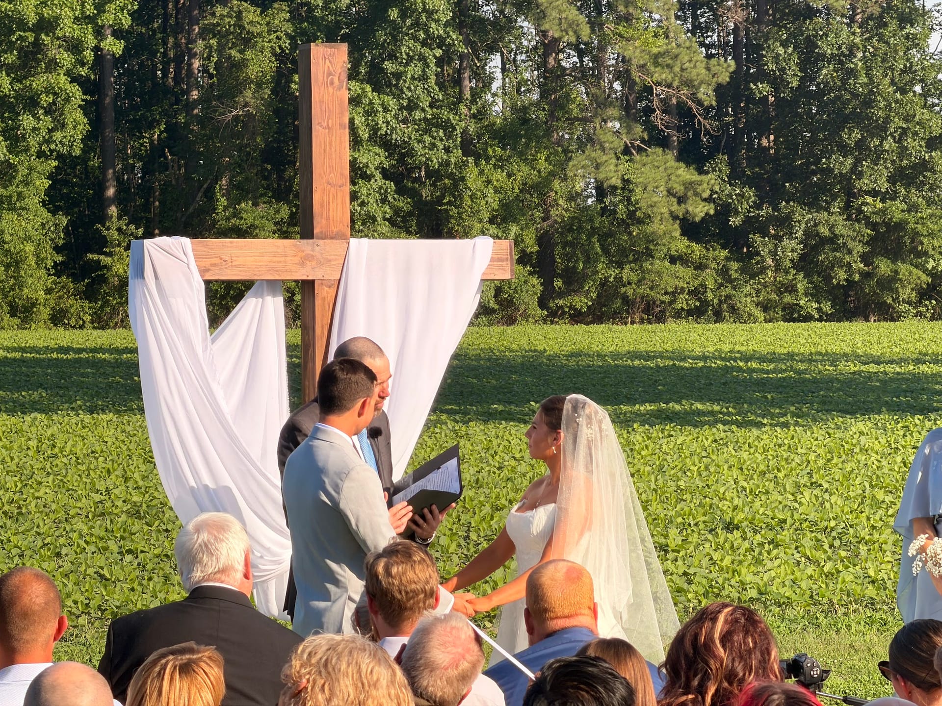 Bride and groom exchange vows beneath a large wooden cross adorned with white fabric on the ceremony lawn at Scarborough House, surrounded by lush green fields and tall trees in the golden evening light.