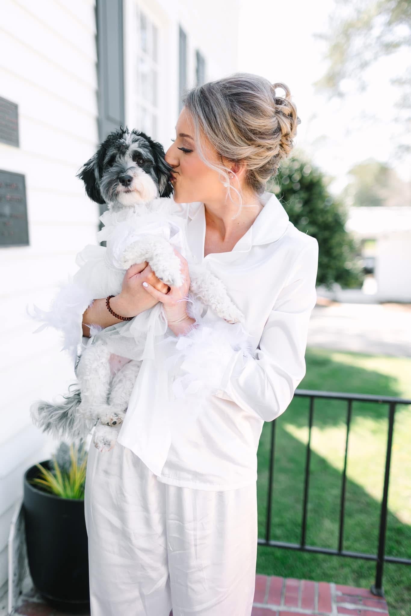 Bride in white getting-ready attire kisses her small black and white dog dressed in a white tulle outfit on the front porch of the historic Scarborough House estate.