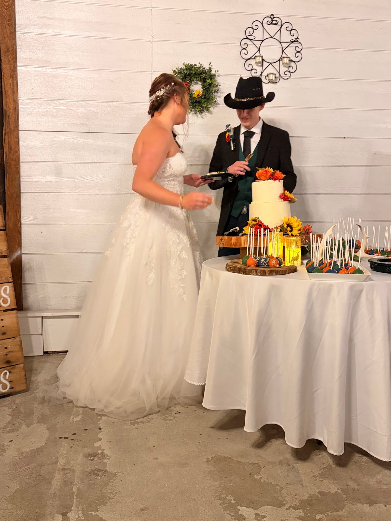 Bride in a white lace ballgown and groom in a black suit and cowboy hat cut their two-tiered wedding cake adorned with sunflowers and autumn blooms at an indoor barn-style reception venue.