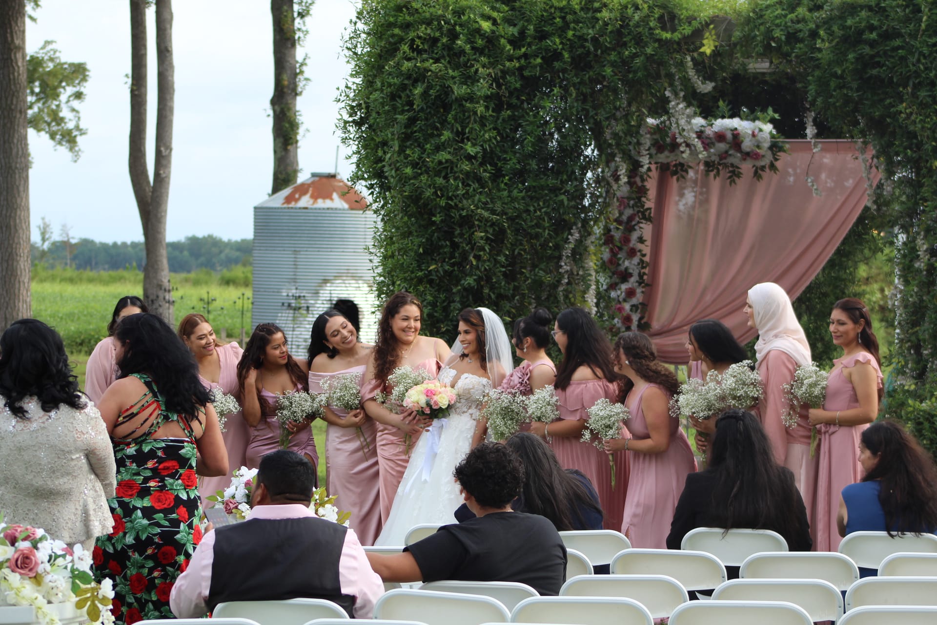 Bride in white gown and veil laughing with bridesmaids in dusty-rose dresses holding baby's breath bouquets beneath a lush greenery arch with pink draping at an outdoor estate wedding ceremony.