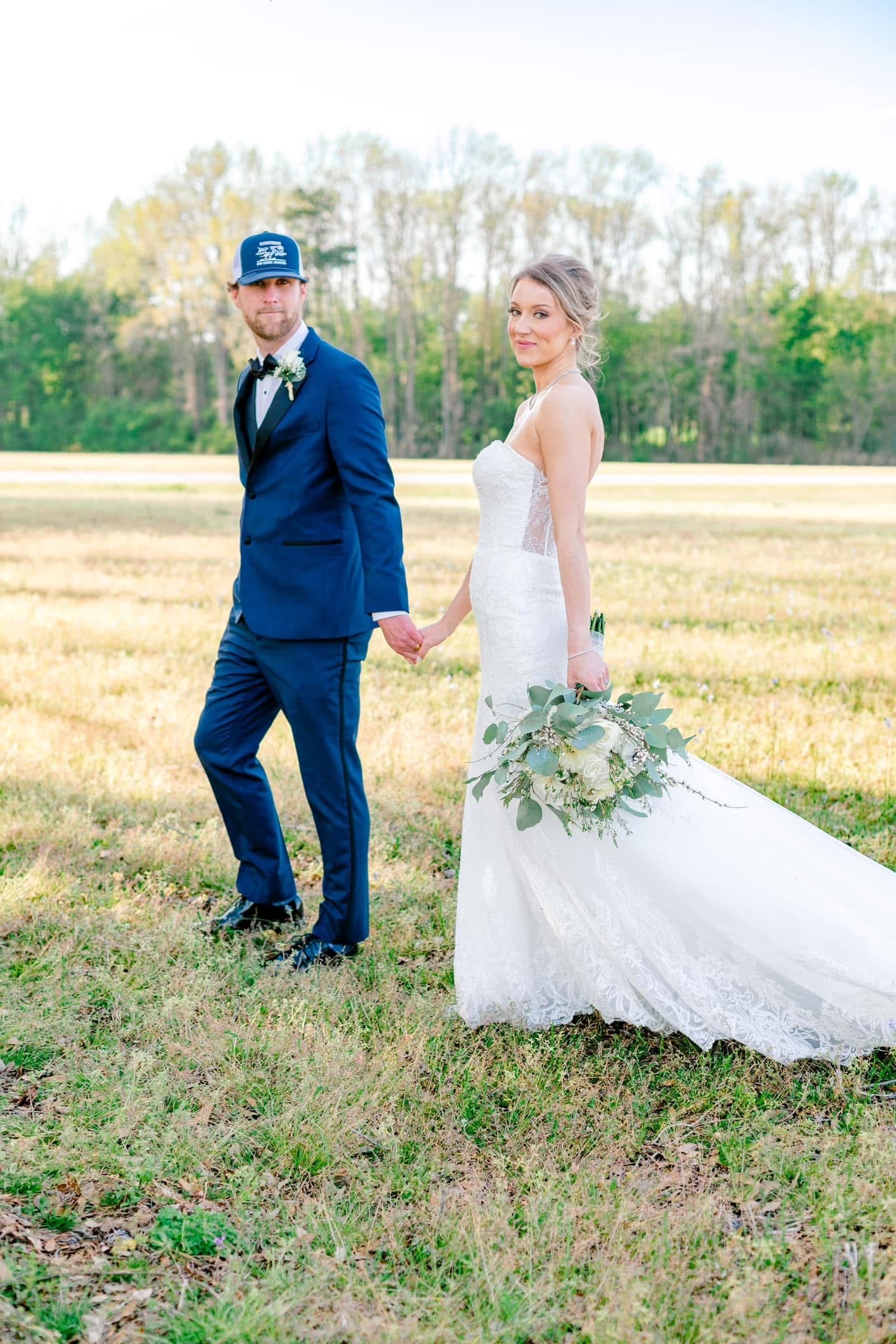 Bride in a lace mermaid gown holding a greenery bouquet looks back over her shoulder while her groom in a navy suit and trucker cap holds her hand as they walk through the golden farmland fields at Scarborough House.
