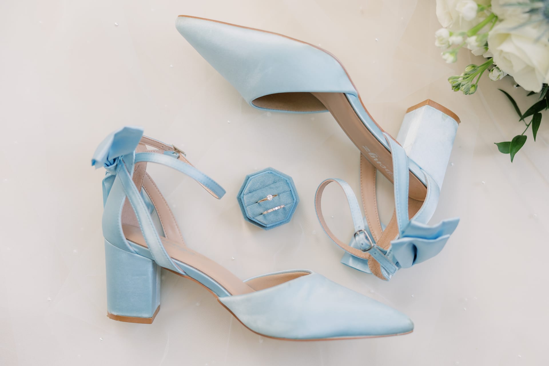 Light blue satin block-heel wedding shoes with bow ankle straps arranged around a blue velvet ring box holding two wedding bands, with white florals in the background.