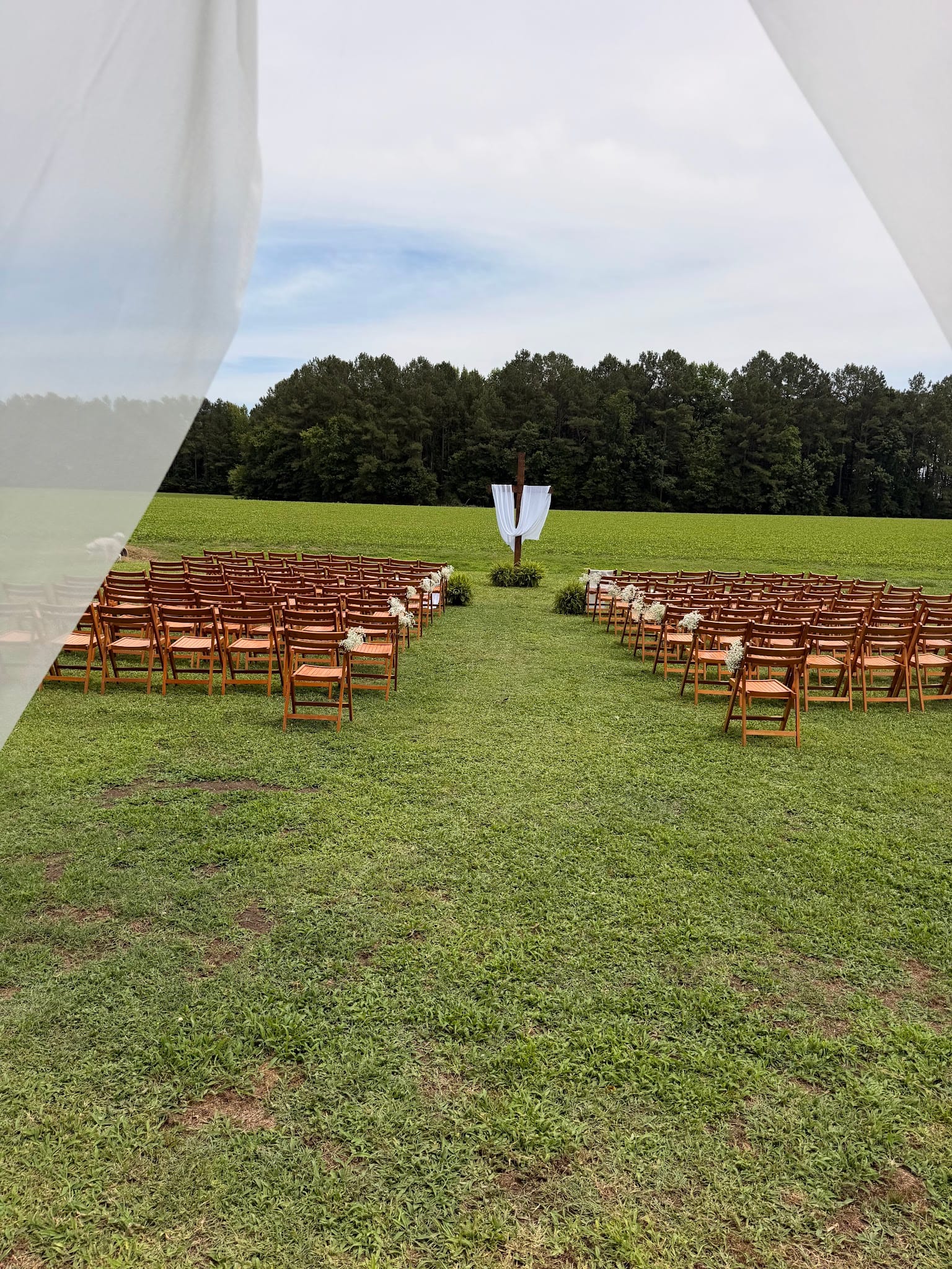 Outdoor wedding ceremony setup on a green lawn with rows of wooden folding chairs flanking a grass aisle leading to a rustic cross altar draped in white fabric, surrounded by open farmland and a treeline backdrop at Scarborough House.