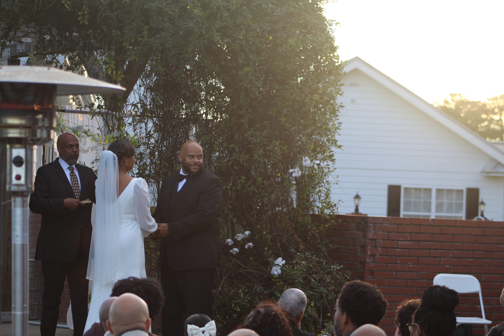 Groom smiles warmly at his bride during an outdoor evening wedding ceremony at a historic estate garden, with soft golden-hour light glowing behind magnolia-covered greenery.