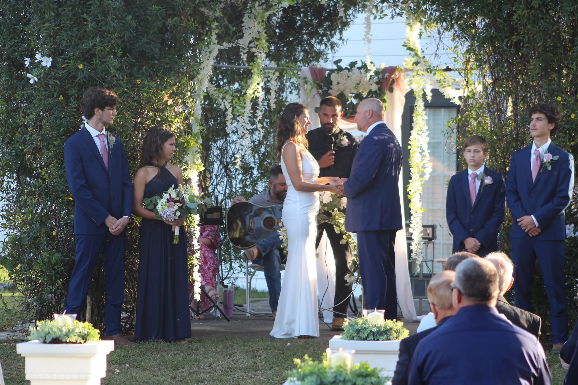 Bride and groom exchange vows beneath a lush floral arch on the ceremony lawn at Scarborough House, flanked by their wedding party in navy and blush.