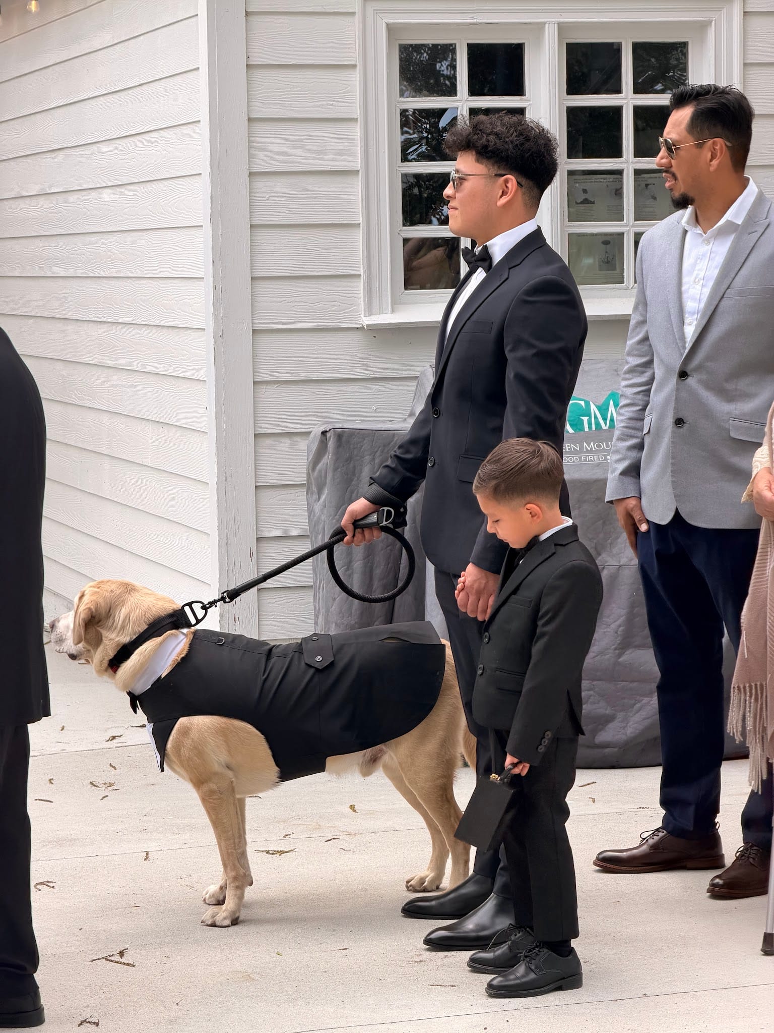 A groomsman in a black tuxedo leads a golden dog dressed in a matching tuxedo jacket while a young boy in a suit stands beside them outside a white historic estate wedding venue.