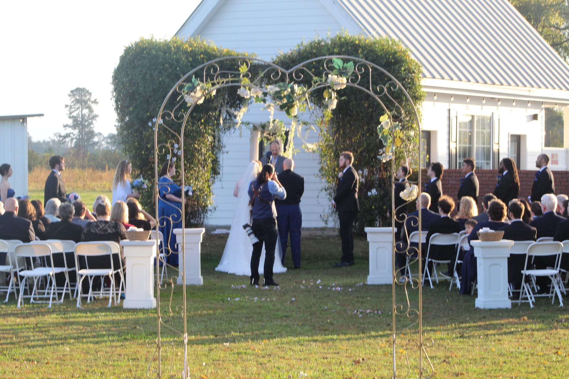 Wedding ceremony on the lawn at Scarborough House viewed through an ornate gold arch adorned with white flowers, with guests seated on either side and a white farmhouse building in the background.