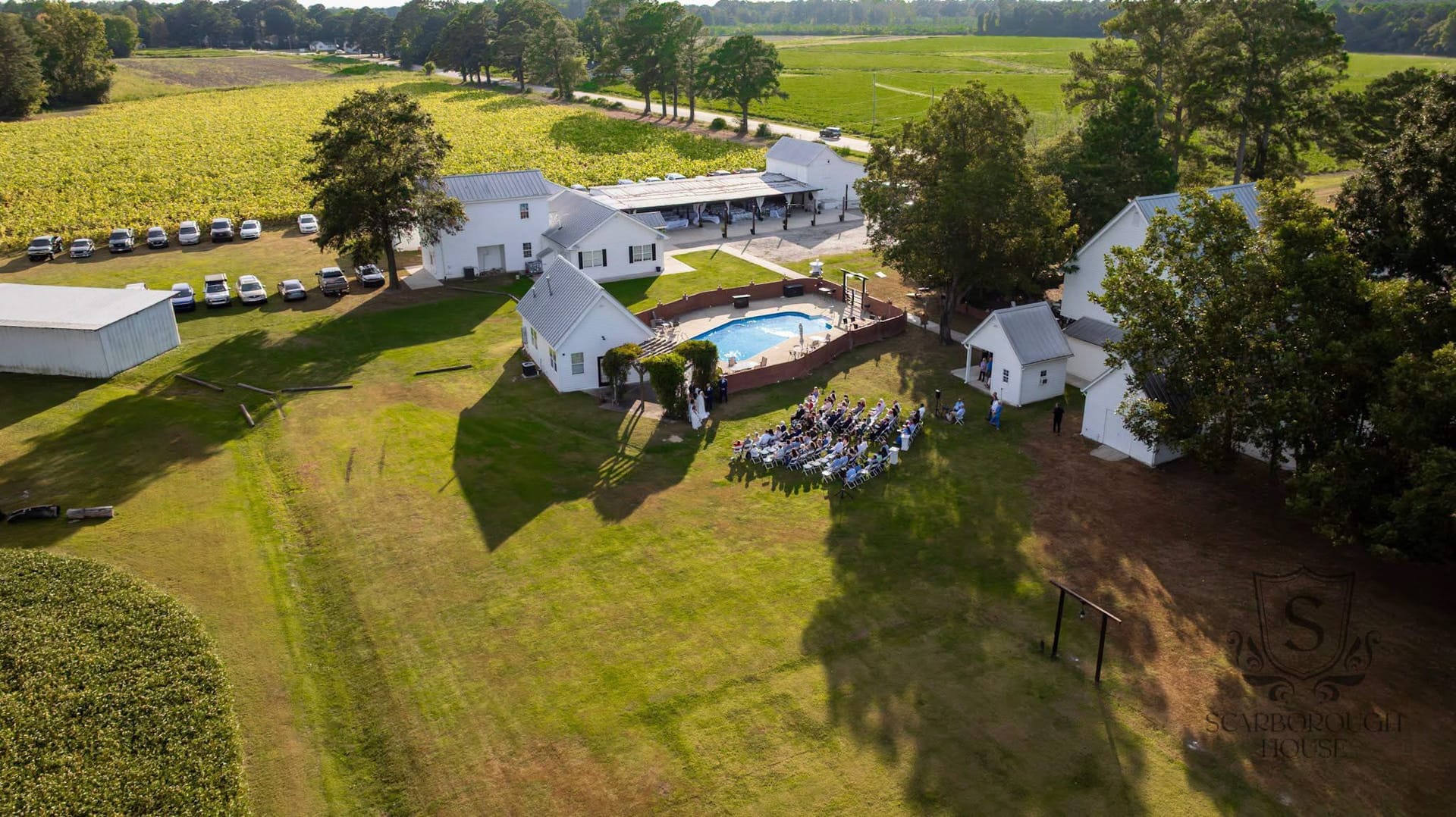 Aerial drone view of a wedding ceremony in progress on the lawn at Scarborough House historic estate, with white manor buildings, a swimming pool, oak trees, and expansive farmland fields surrounding the property.