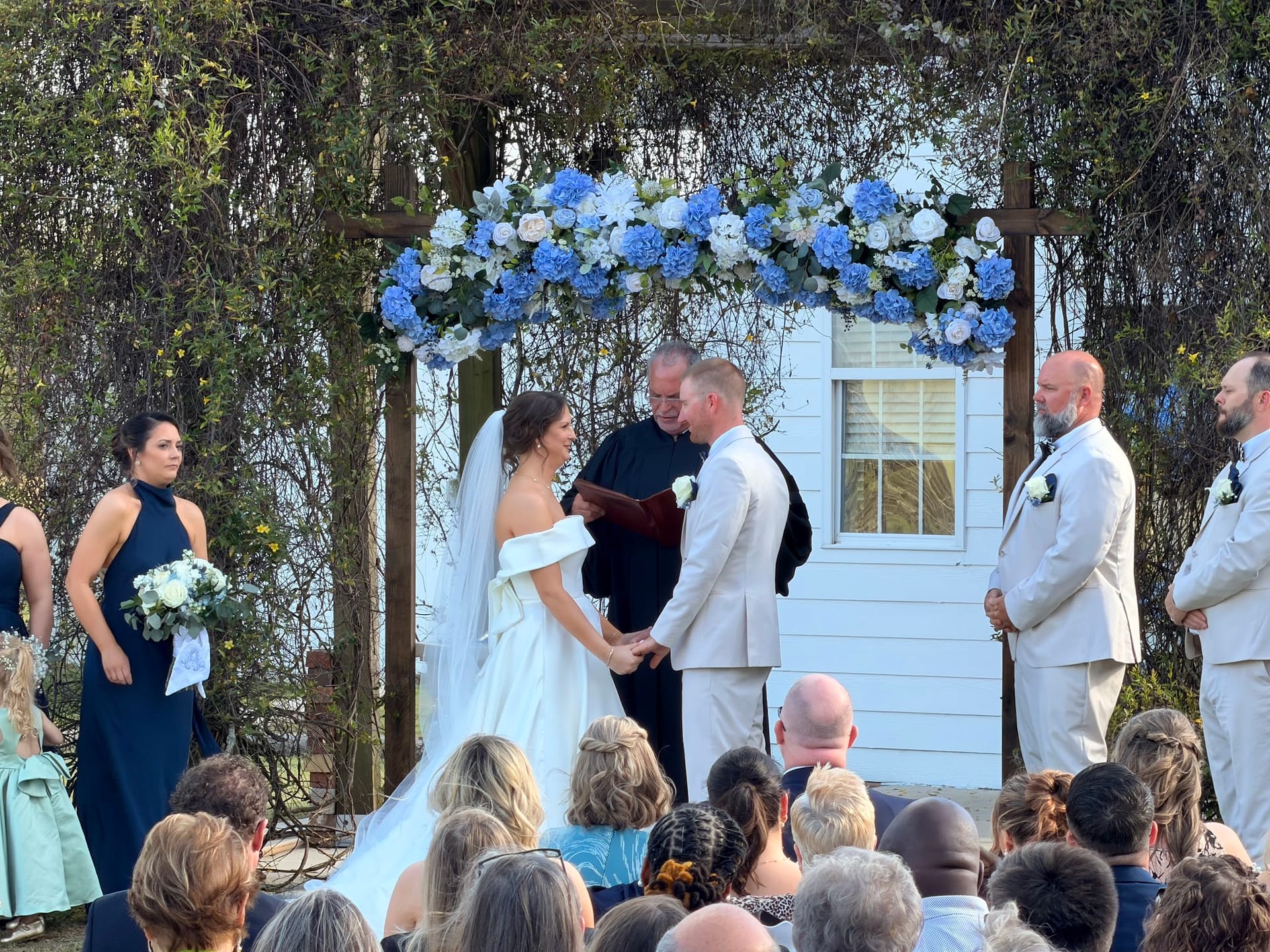 Bride and groom exchange vows beneath a rustic pergola adorned with blue hydrangeas and white roses on the ceremony lawn at Scarborough House, surrounded by wedding party and seated guests.