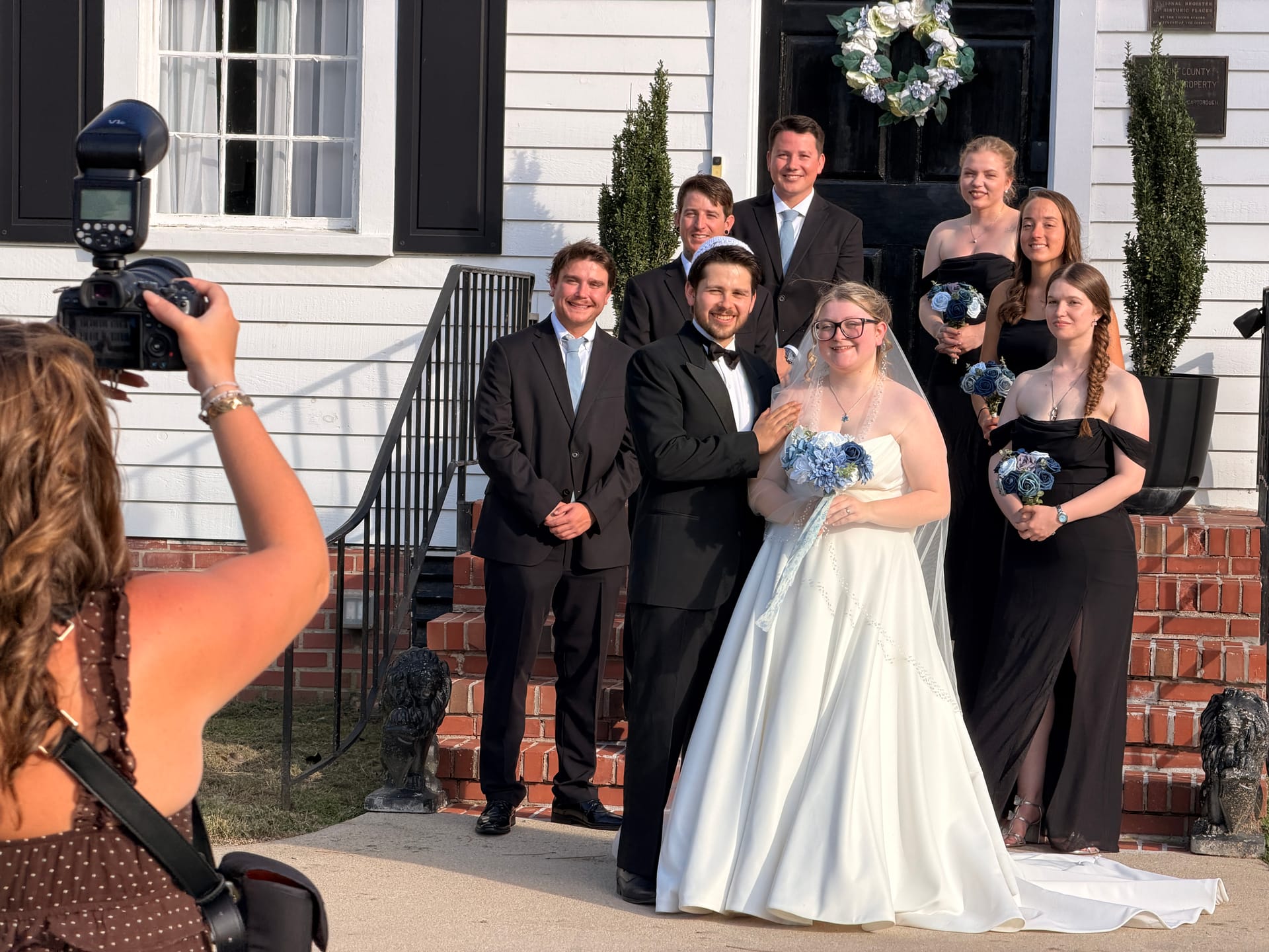 Bride in a white ballgown and groom in a black tuxedo pose with their wedding party in black attire holding blue bouquets on the front steps of the 1821 Scarborough House manor while a photographer captures the moment.