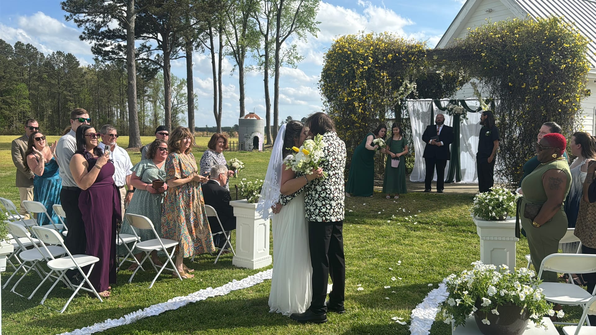 Bride and groom share a kiss at an outdoor ceremony on the sunlit lawn at Scarborough House, surrounded by applauding guests and framed by a lush floral arch with white draping.