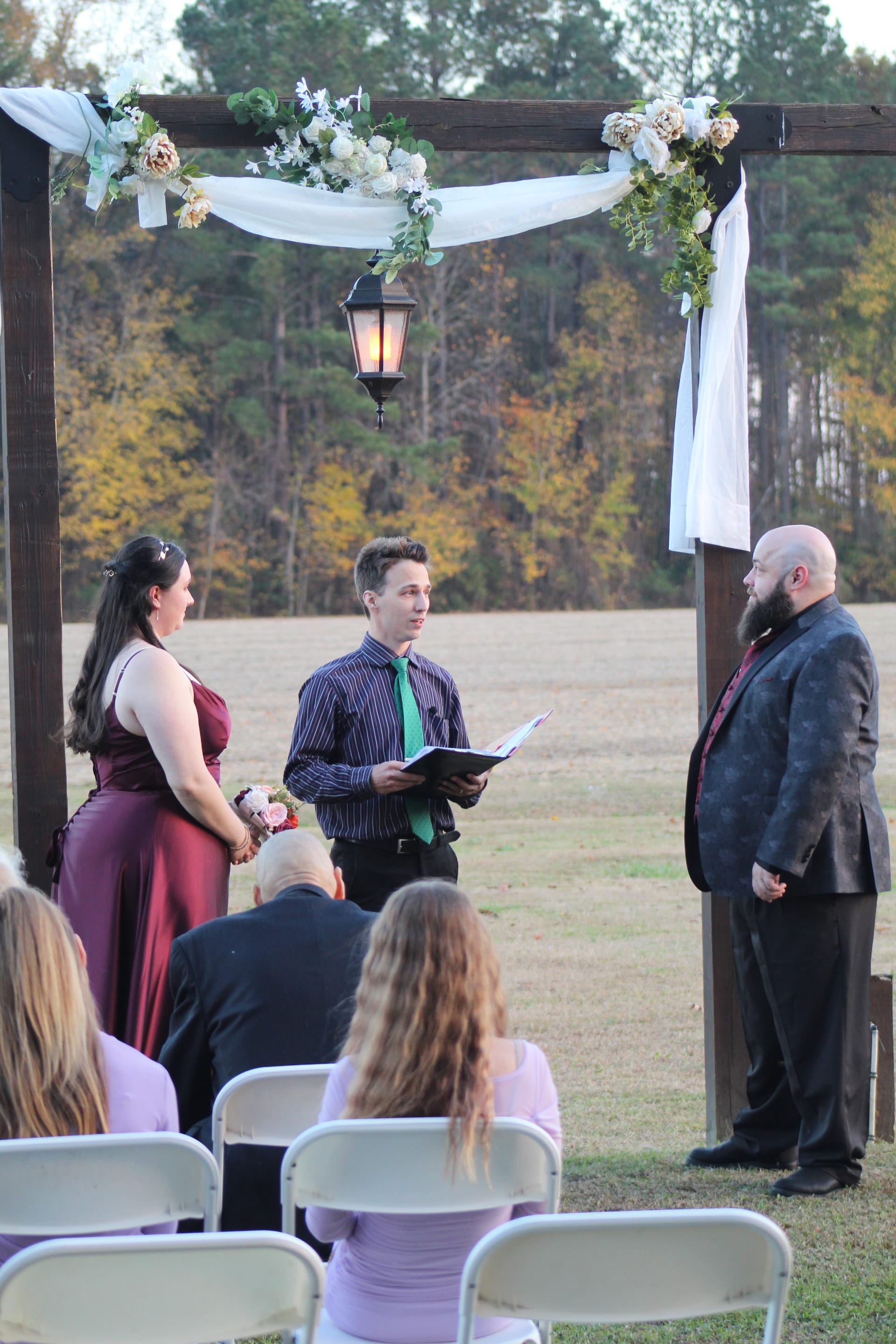 A bride in a burgundy gown and groom in a dark suit exchange vows beneath a wood arch adorned with white flowers and draped fabric on the ceremony lawn at Scarborough House, surrounded by autumn trees and open farmland fields.