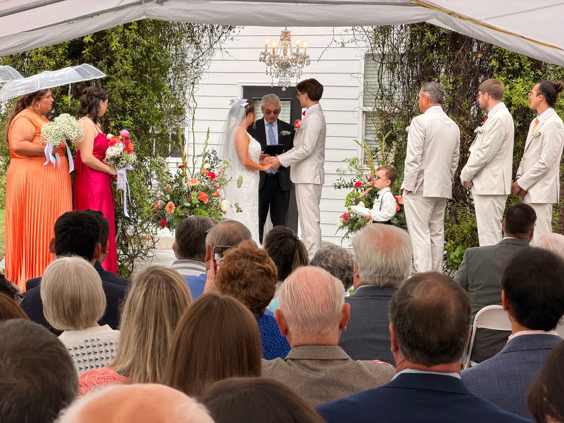 Bride and groom exchange vows beneath a chandelier-lit tent surrounded by colorful floral arrangements and their wedding party at a historic outdoor estate ceremony.