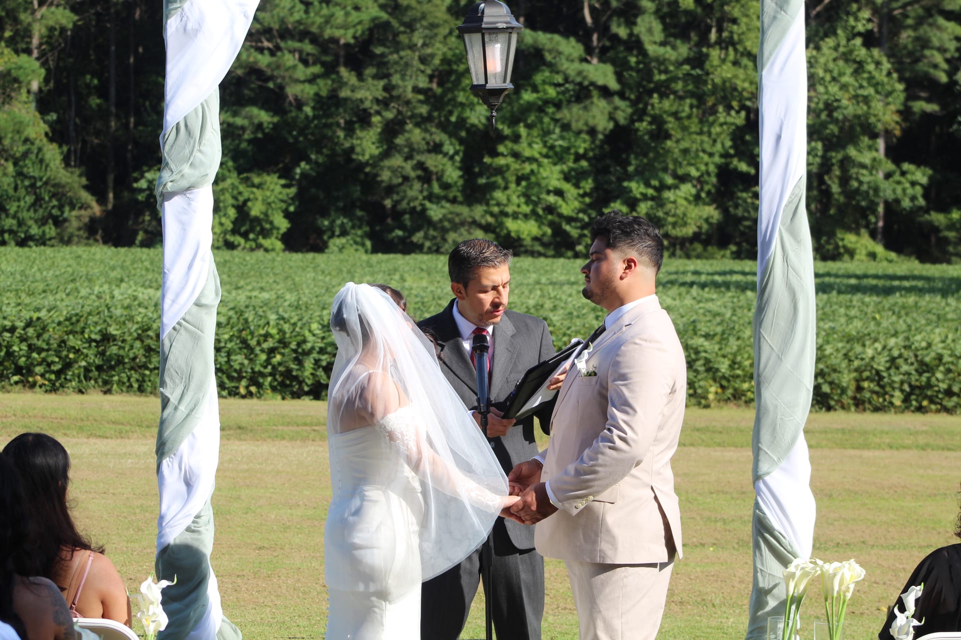 Bride and groom hold hands while exchanging vows beneath a draped arch on the ceremony lawn at Scarborough House, with green farmland fields and trees in the background.