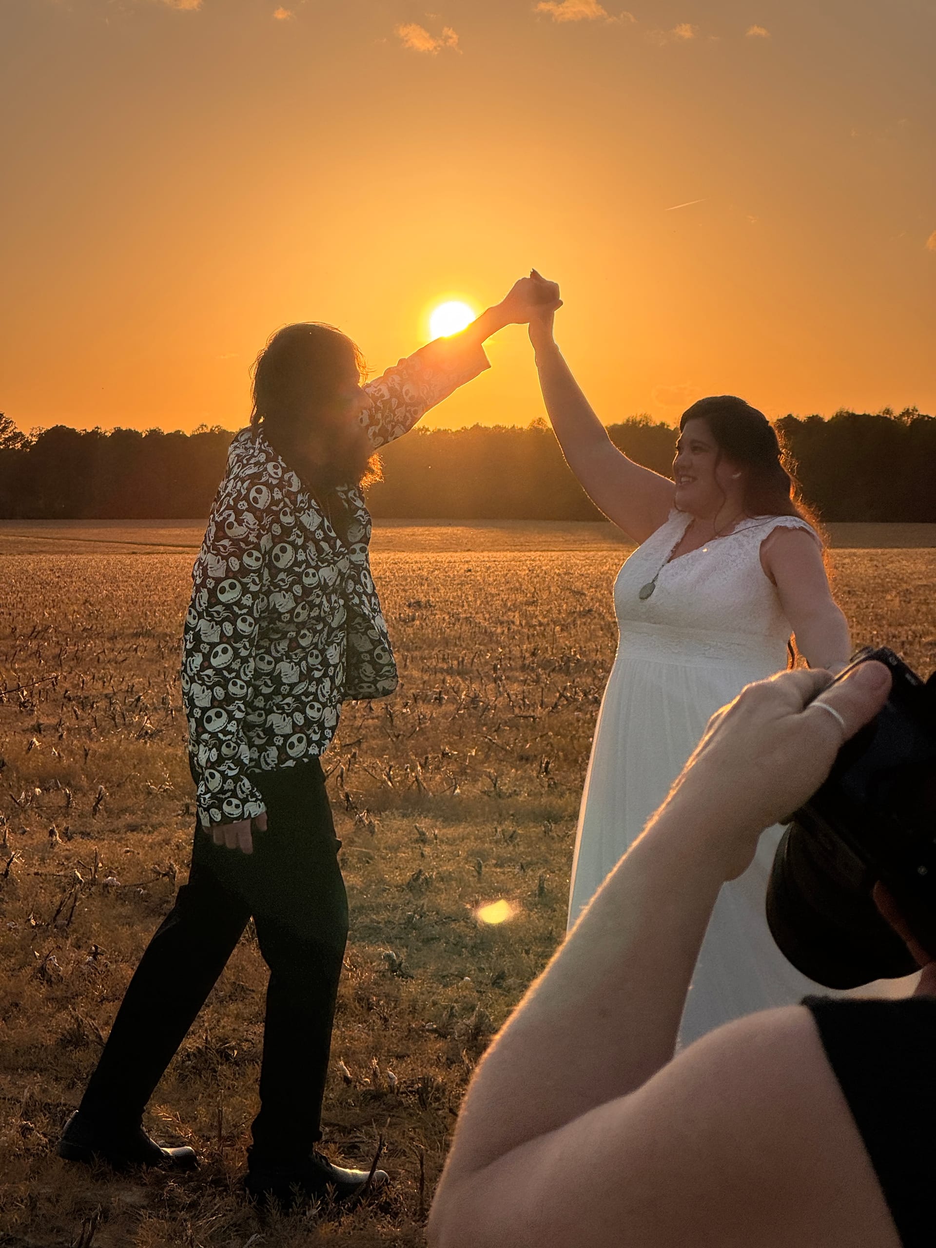 Bride in white gown and groom in a patterned shirt dance together hand-in-hand in an open farmland field at golden sunset on their wedding day.