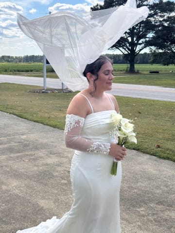 Bride in a fitted white off-shoulder gown holding a white rose bouquet as her veil billows dramatically in the wind on the ceremony lawn of Scarborough House estate.
