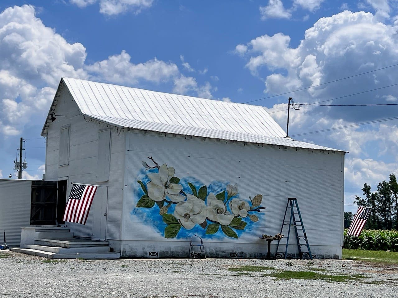 A white estate barn with a painted magnolia mural and American flags under a bright blue sky