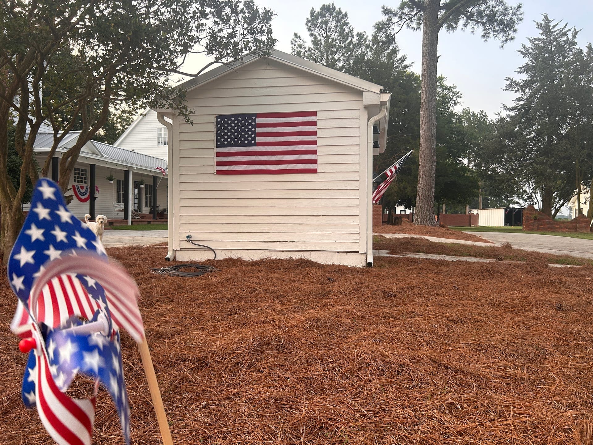 A white estate outbuilding flying a large American flag with patriotic pinwheels in the foreground and a dog on the lawn