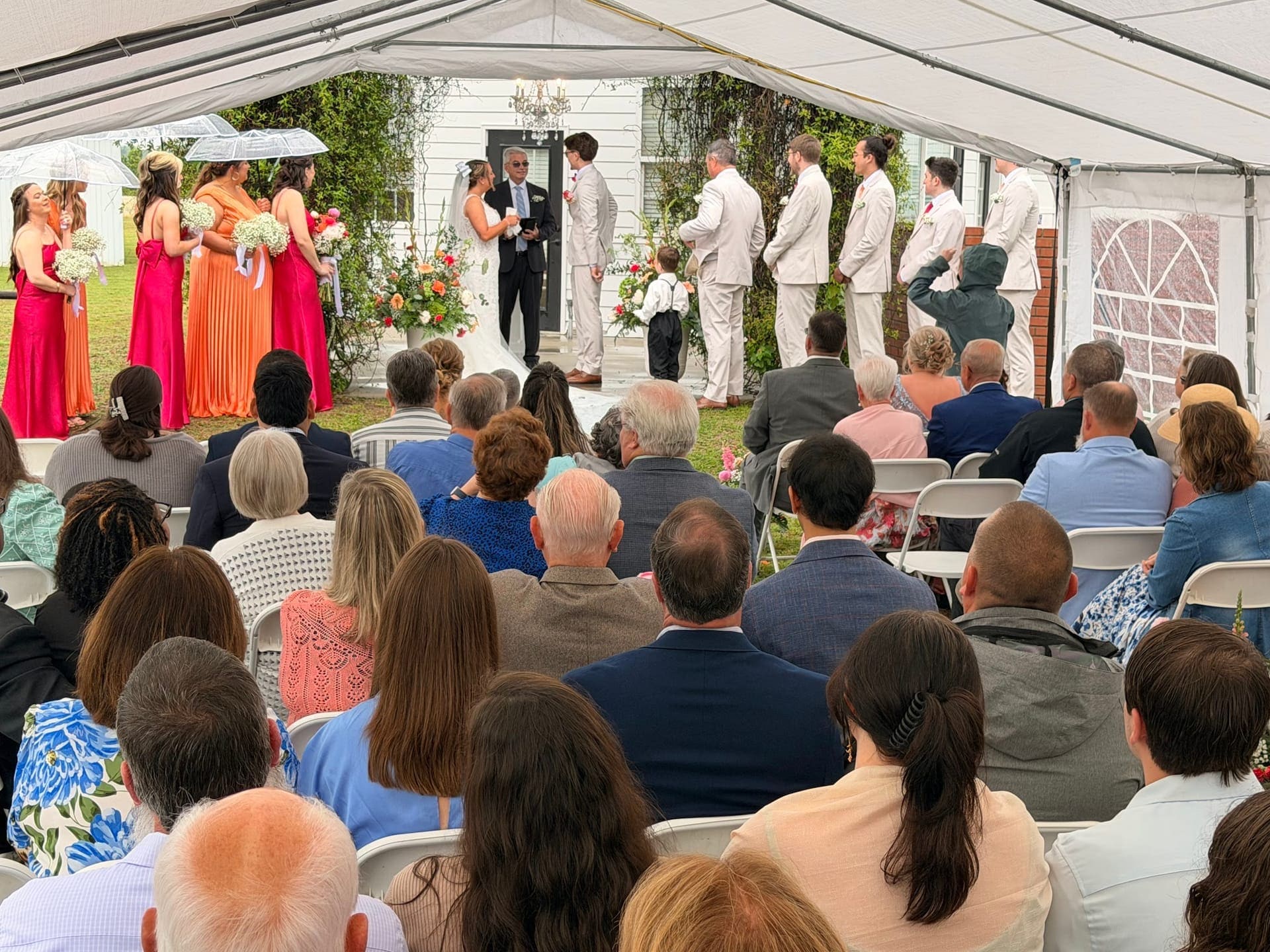 Guests seated under a white tent watch a bride and groom exchange vows at an outdoor garden ceremony with bridesmaids in orange and pink at Scarborough House.