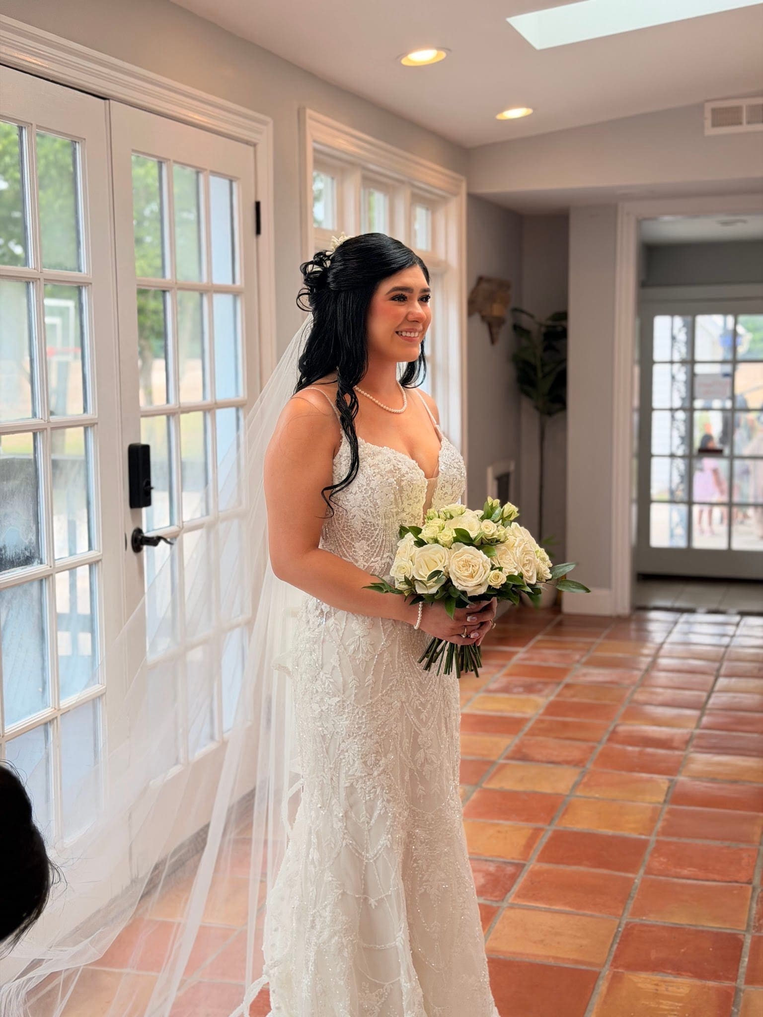 Smiling bride in a beaded lace wedding gown and veil holds a white rose bouquet near the French doors inside Scarborough House.