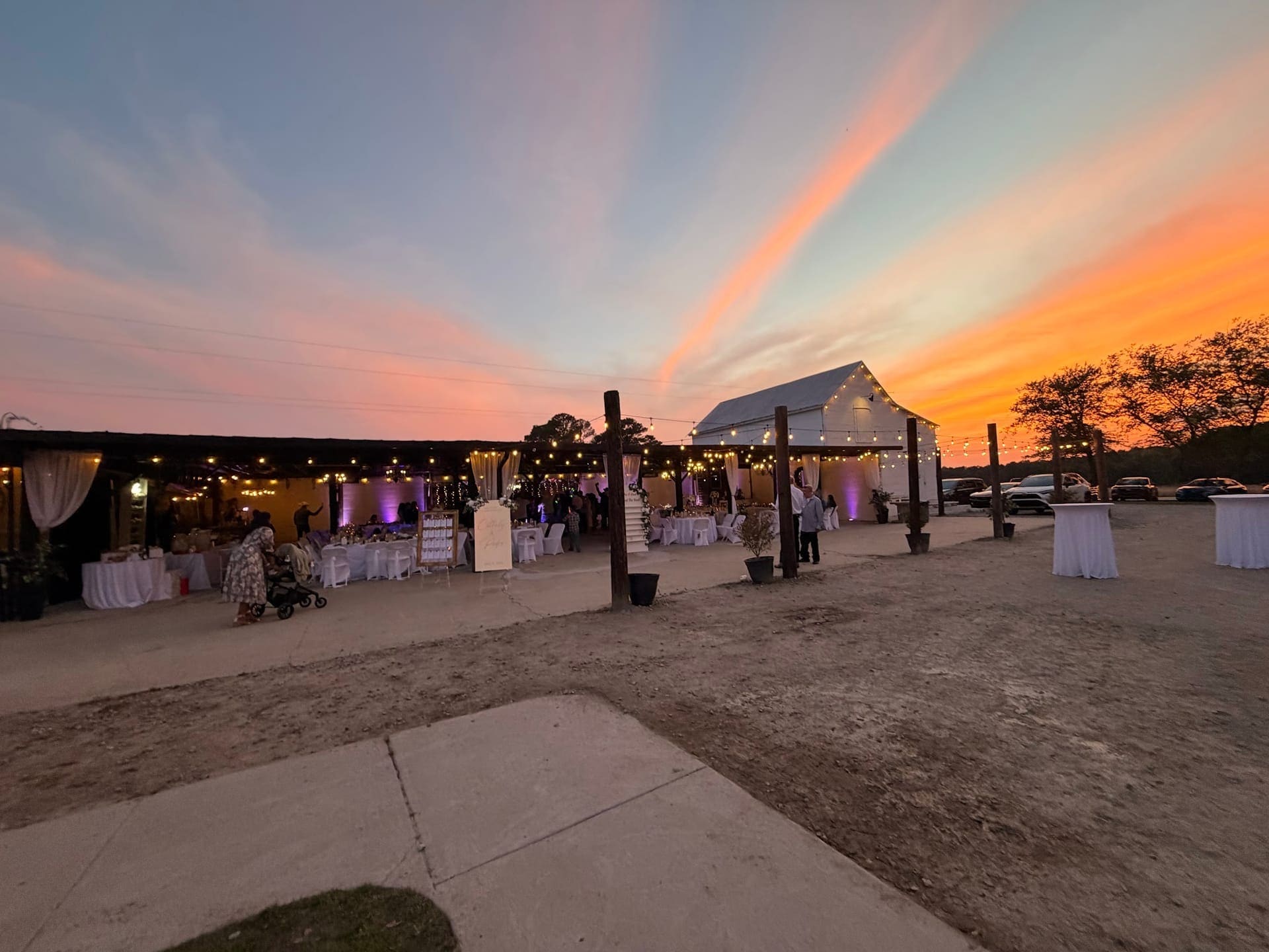 Outdoor wedding reception at Scarborough House glowing with string lights and purple uplighting beneath an orange and pink sunset sky.